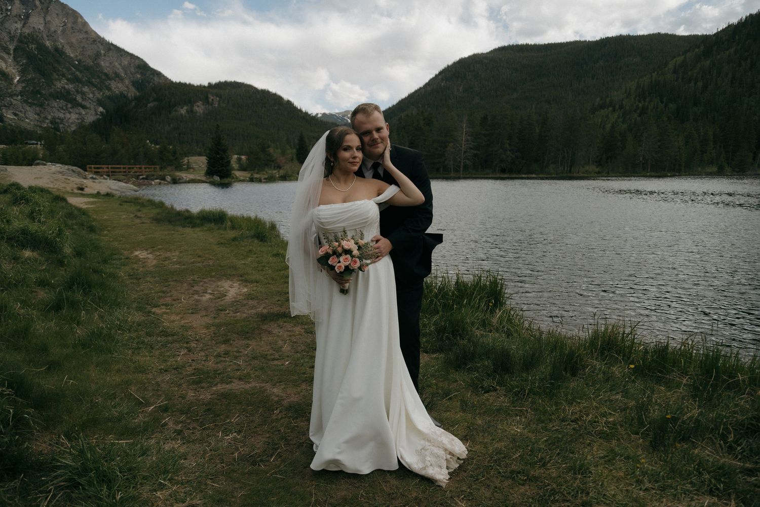 Groom embraces bride holding bouquet near scenic mountain lake during outdoor wedding.
