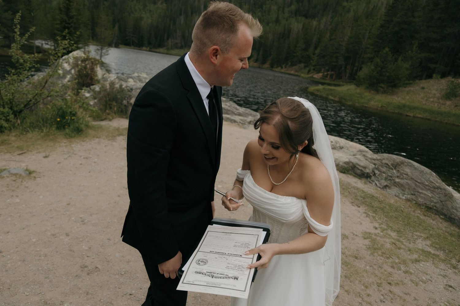 Bride and groom signing wedding certificate outdoors near a mountain lake, wearing formal wedding attire.