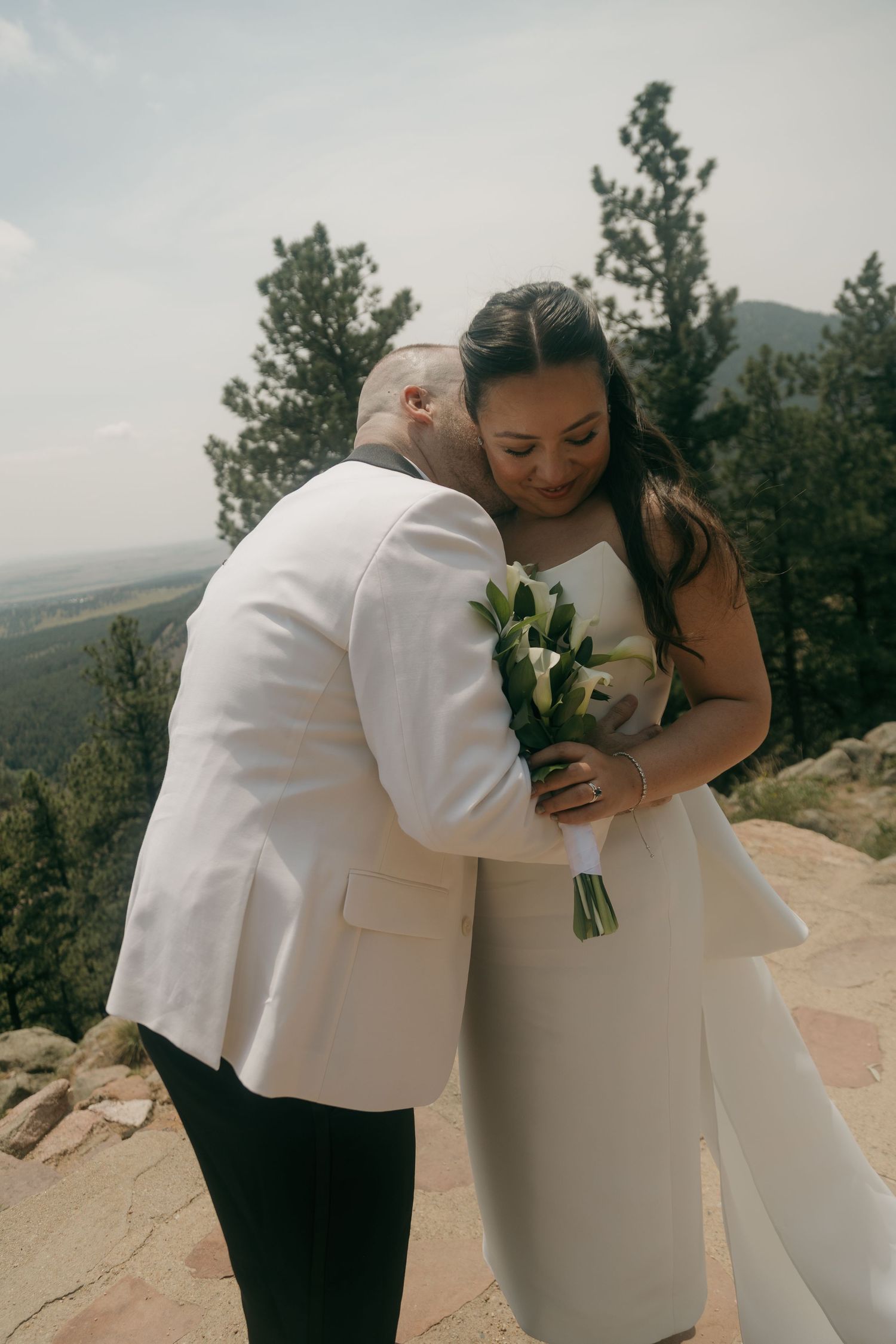 Groom in white jacket kissing bride on the cheek at a scenic mountain wedding location.