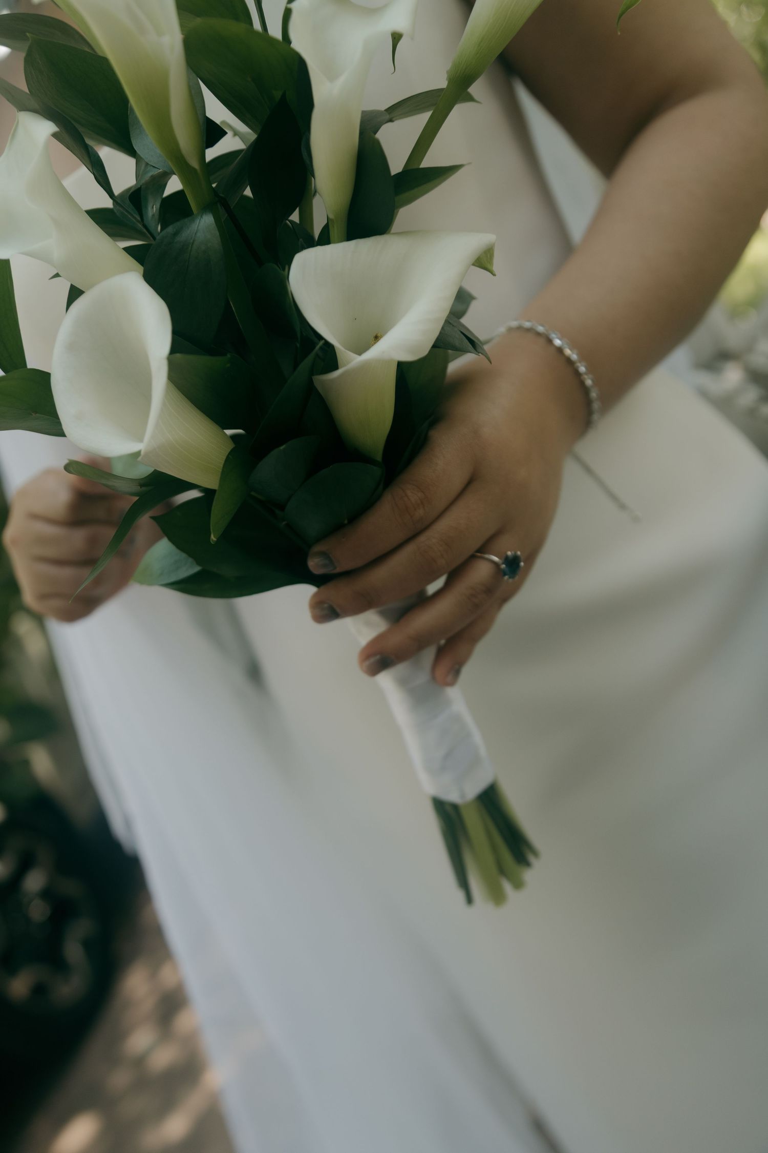 Bride in white gown holds elegant bouquet of white calla lilies with green foliage.