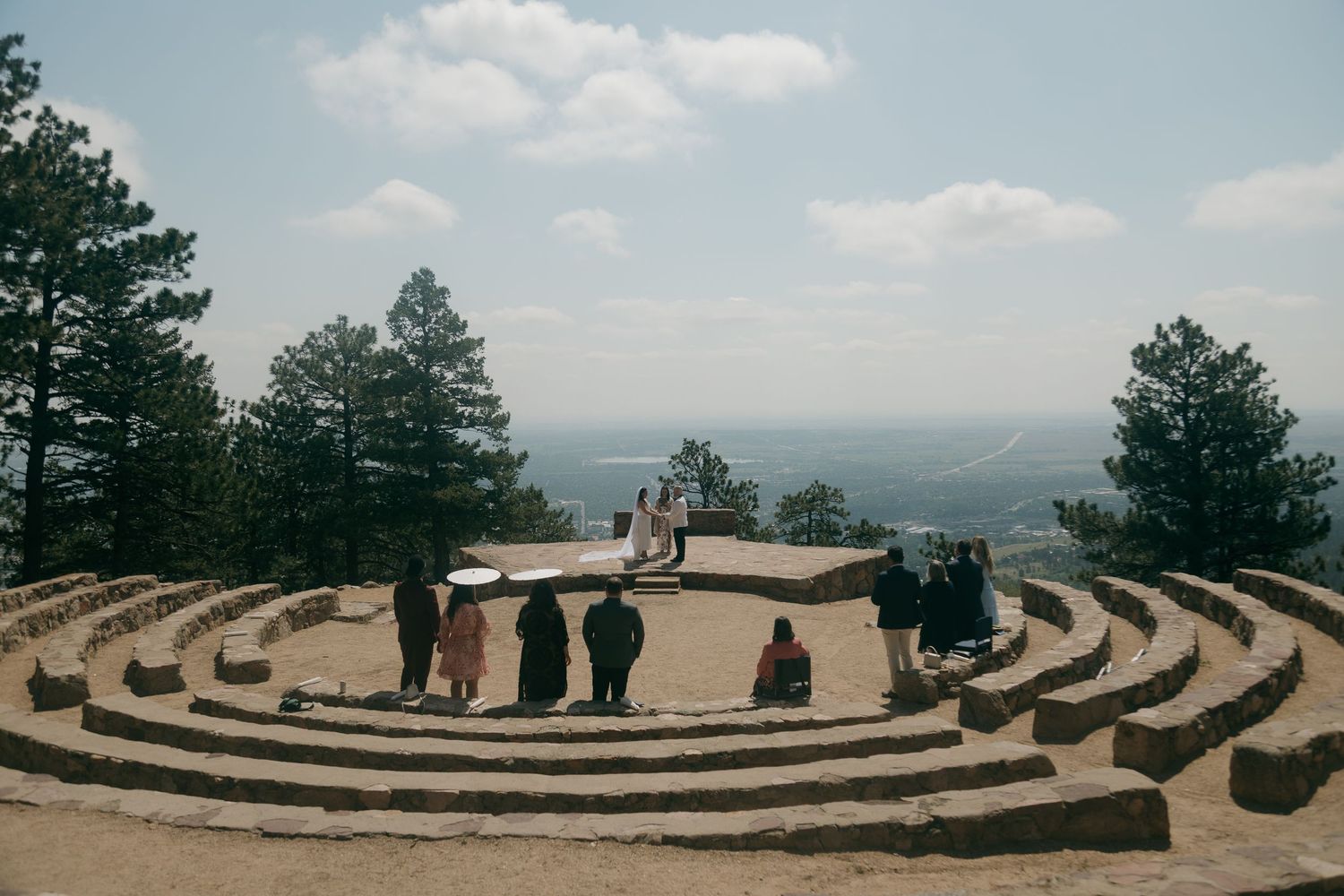 Wedding guests gather in a circular stone amphitheater on a hilltop surrounded by pine trees with a vast landscape view.