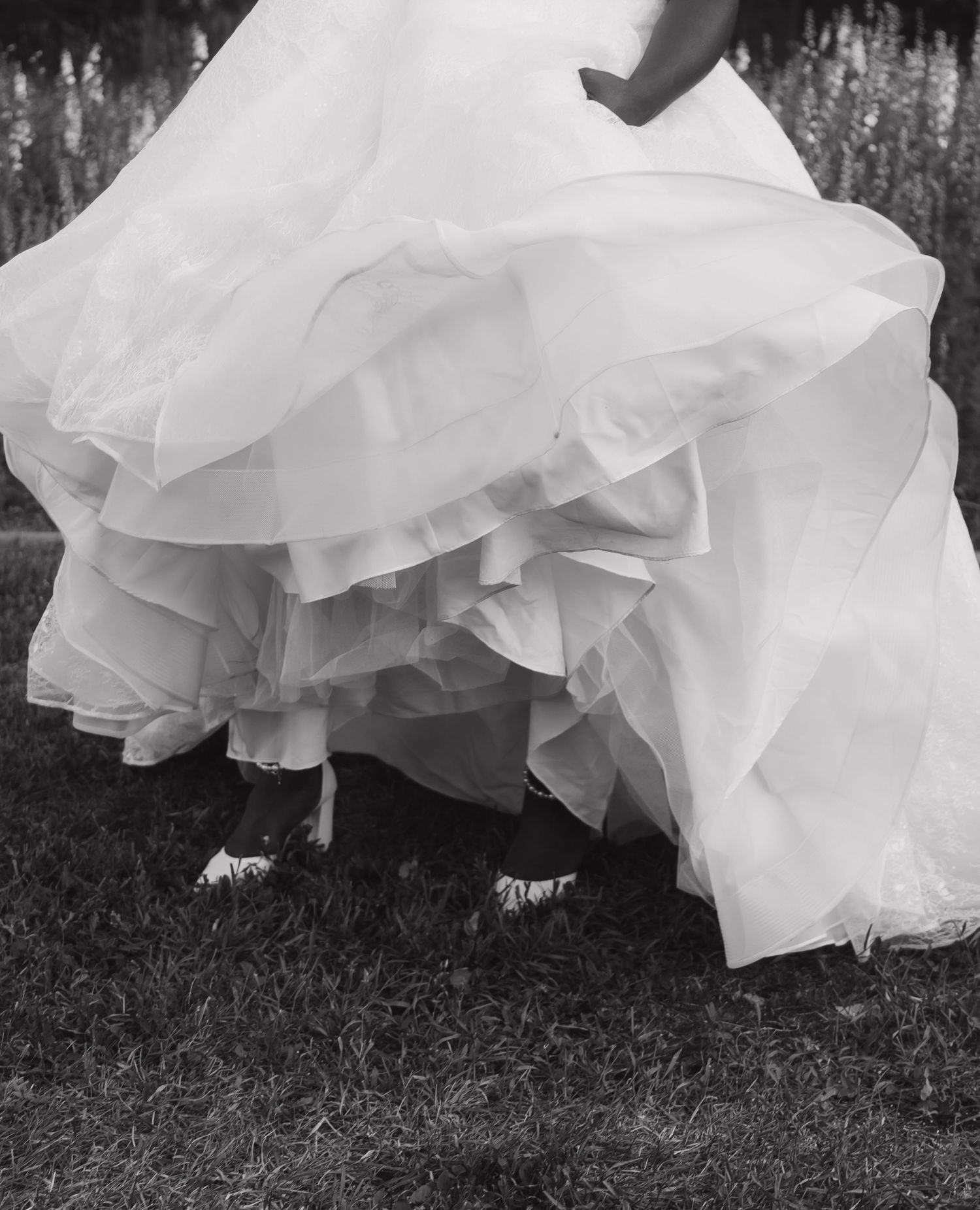 Black and white photo of a bride in a layered ruffled wedding gown standing on grass, showing dress details and shoes.