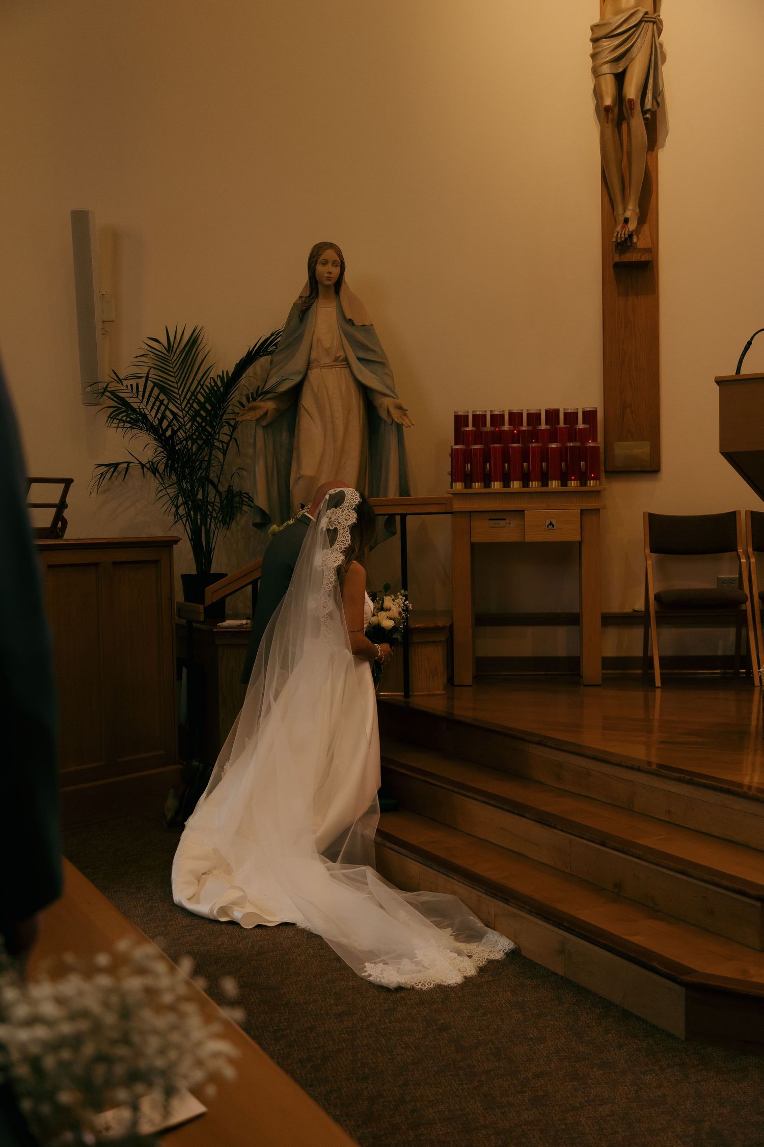 Bride in white gown and veil kneeling before Virgin Mary statue in a warmly lit Catholic church.