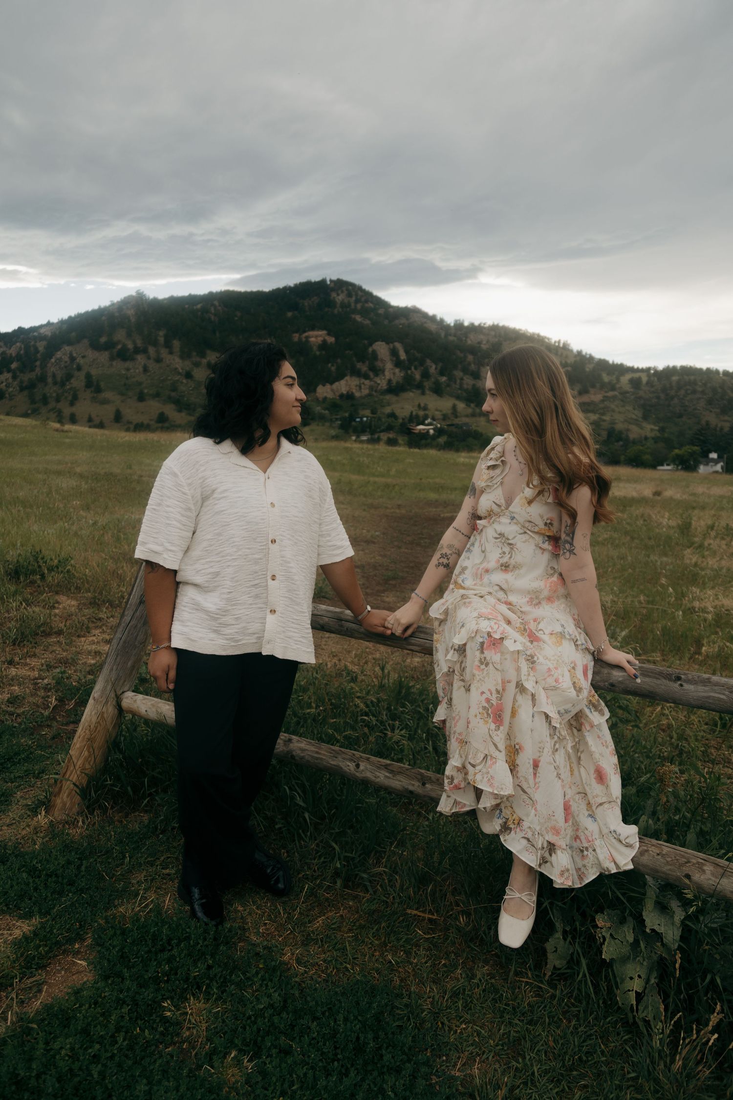 A couple holds hands near a rustic wooden fence with a green mountain landscape behind them.
