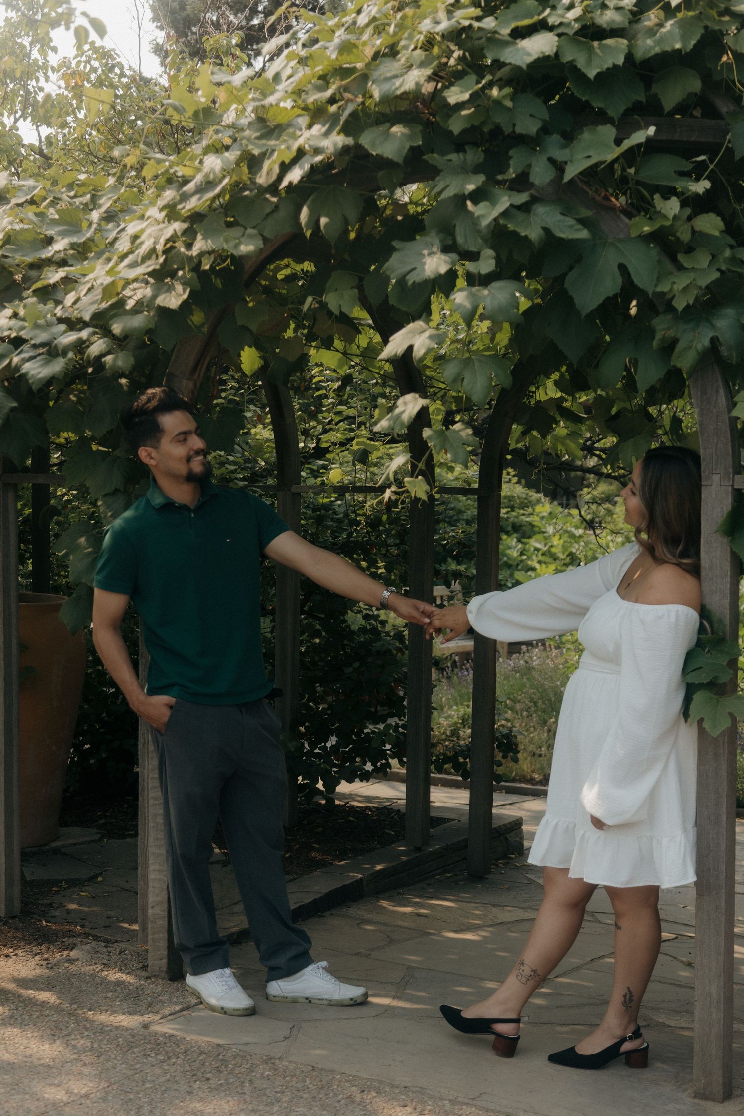 Couple holding hands under a lush green vine-covered pergola in a garden.