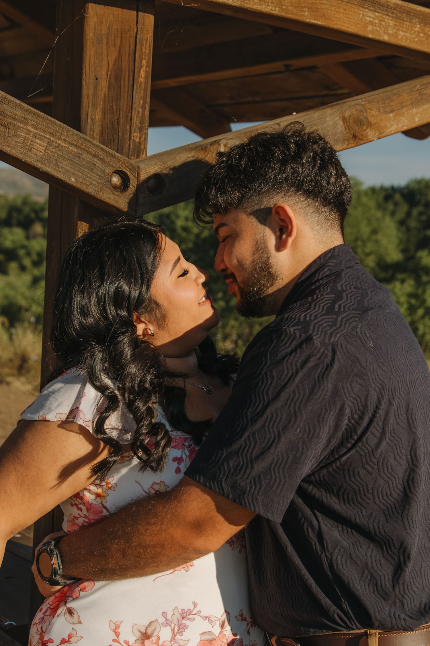 A romantic moment captured between a couple under a wooden structure, with her in a floral dress and him in black.
