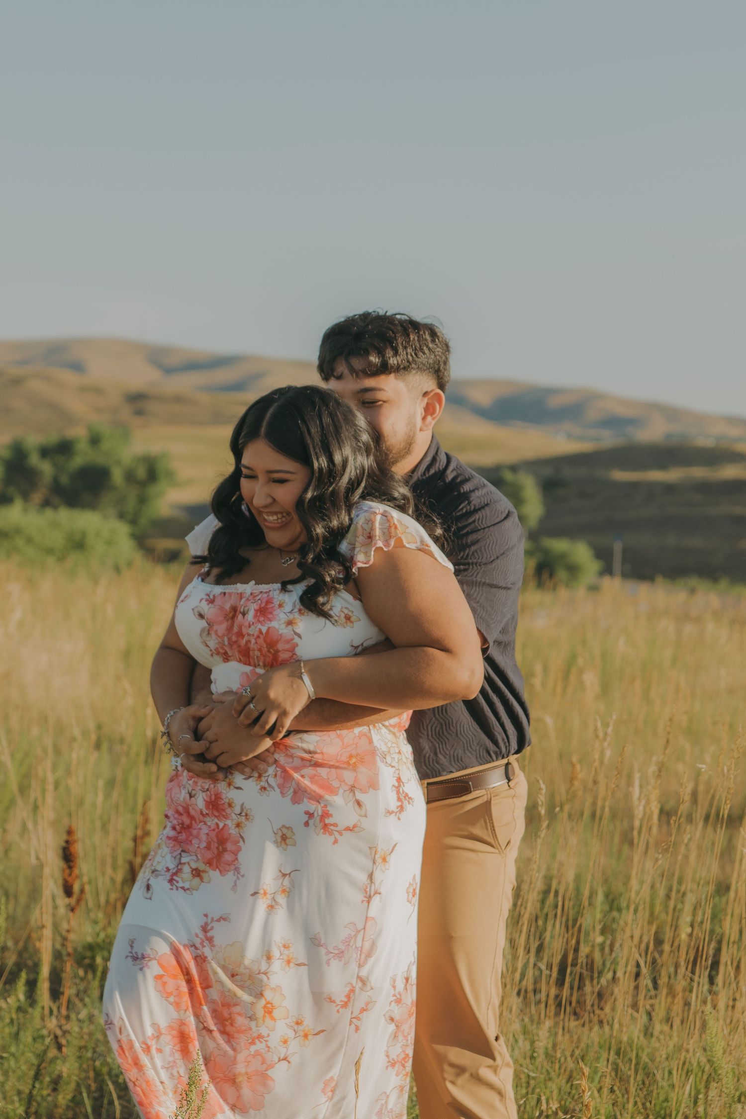 A couple embraces in a scenic field with rolling hills and golden grass in the background.