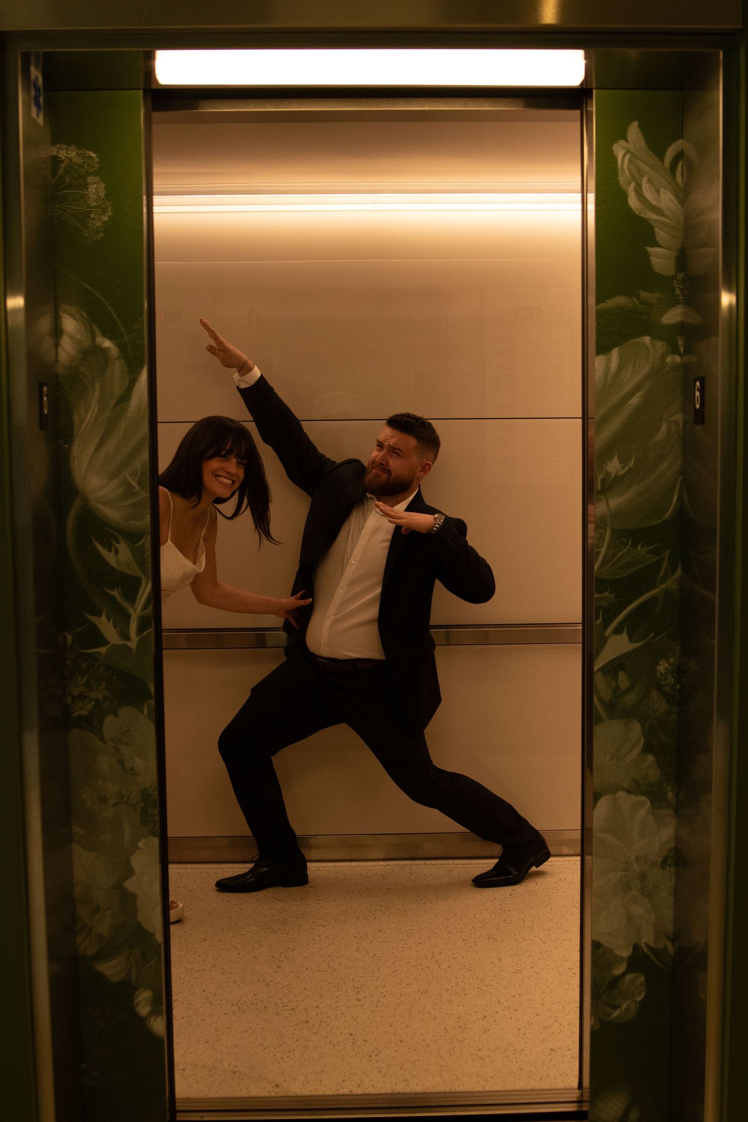 Two people strike dramatic poses in an elevator with reflective walls.