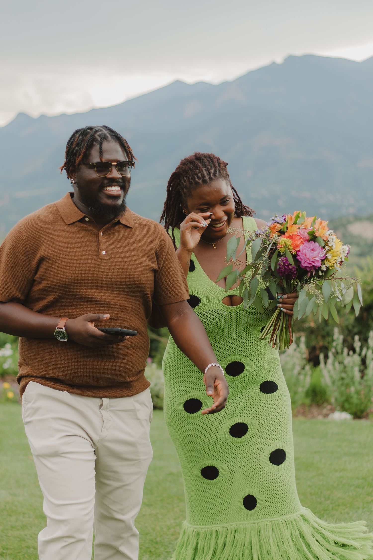 A couple shares a joyful moment in formal attire against a mountain backdrop with colorful flowers.