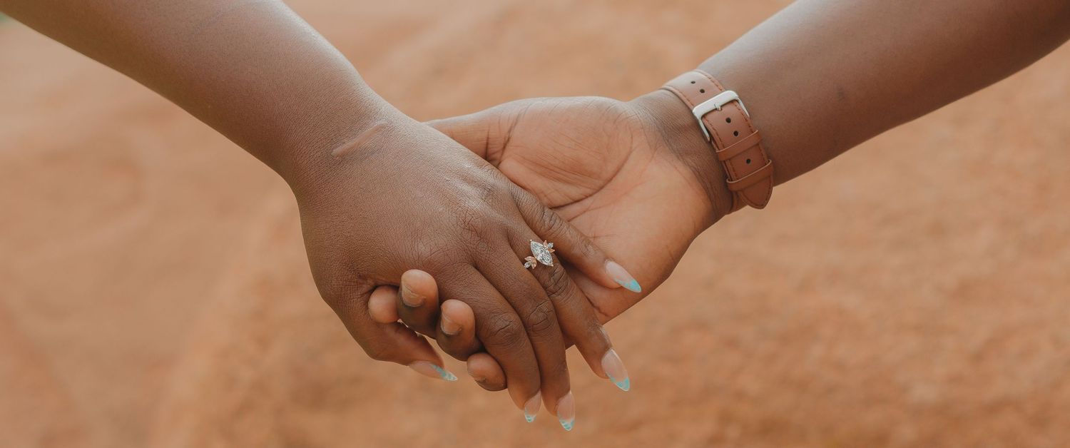 Two hands wearing rings hold each other against a soft brown background, symbolizing love and commitment.