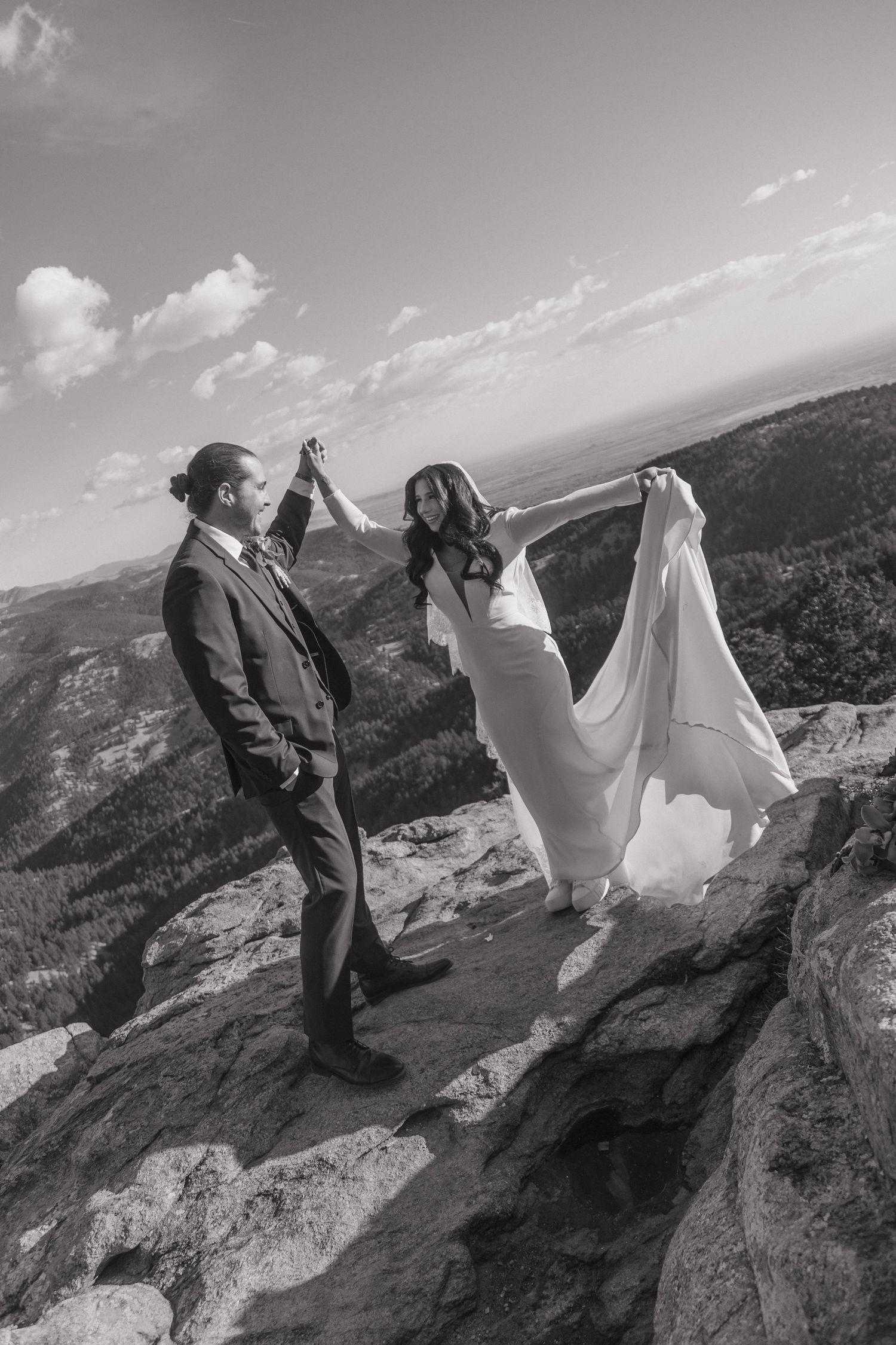 Black and white photo of bride and groom dancing on rocky mountain peak, bridal gown billowing in the breeze.