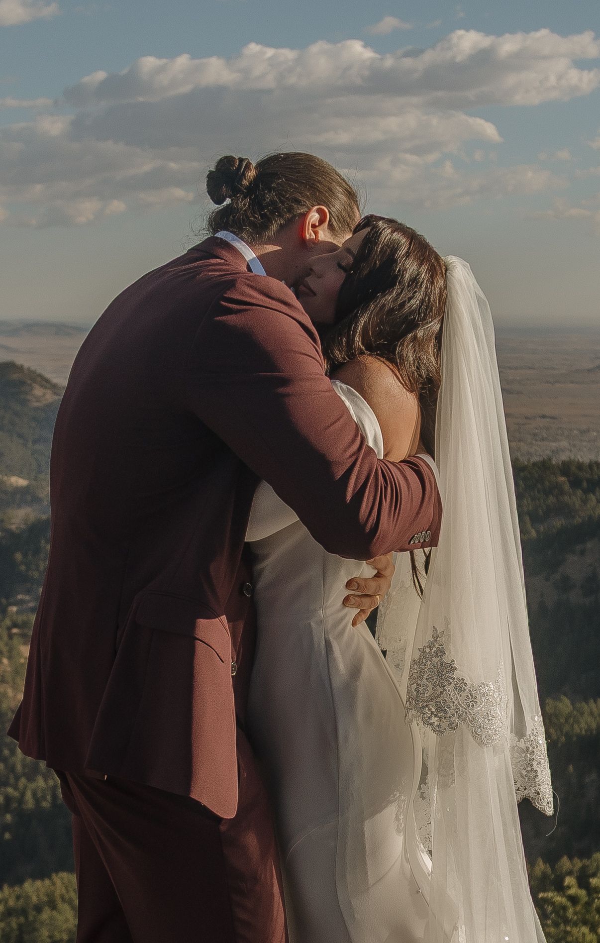 Bride and groom share first kiss in burgundy suit and white veil with mountain views.