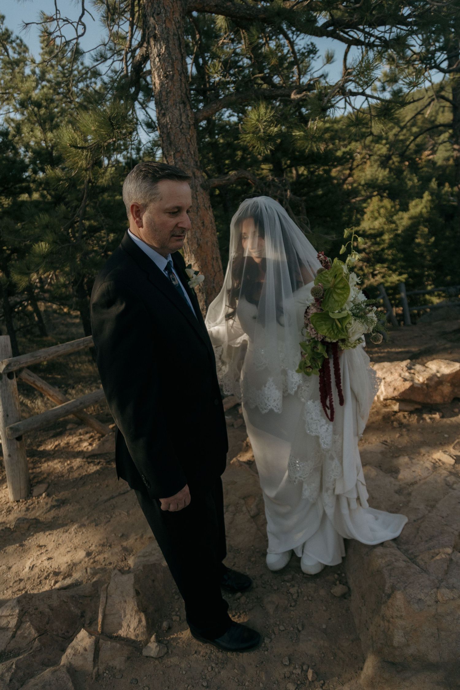 Groom in black suit standing beside veiled bride holding green bouquet during outdoor mountain wedding ceremony.