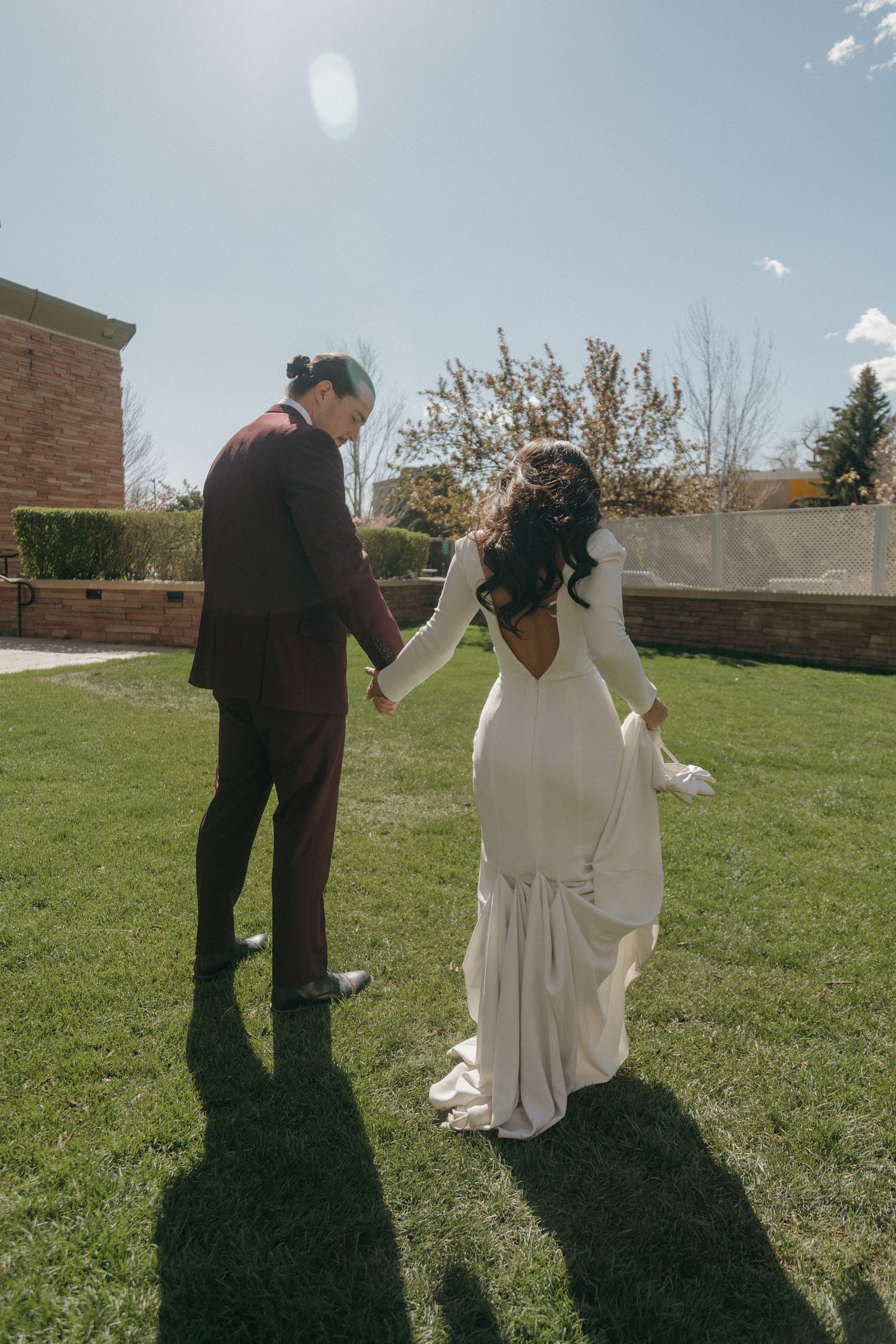 Bride in fitted white long-sleeve gown holding hands with groom in burgundy suit on sunny lawn.