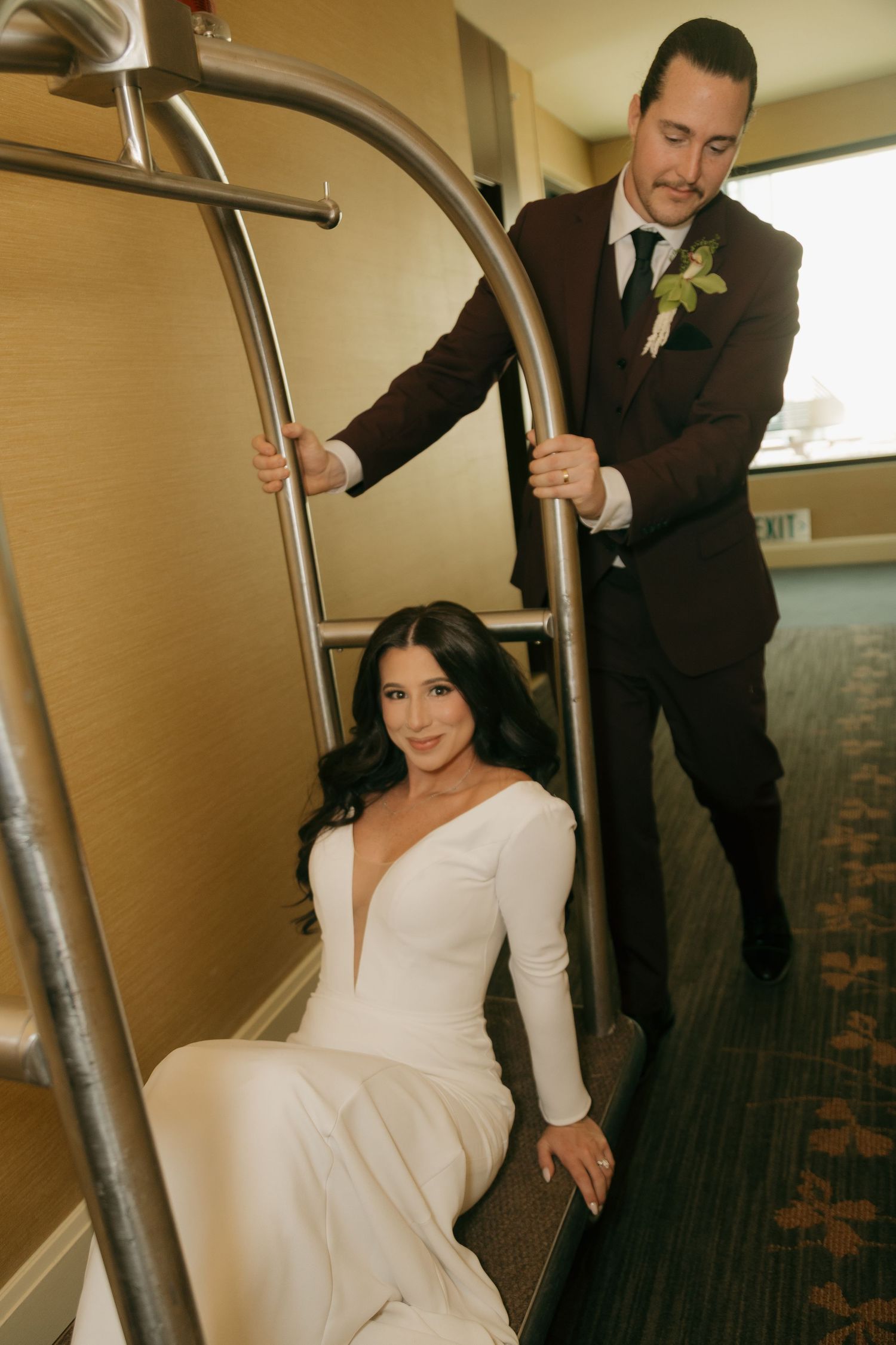 Bride in white gown sitting on a luggage cart while groom in burgundy suit stands behind her in a hotel.