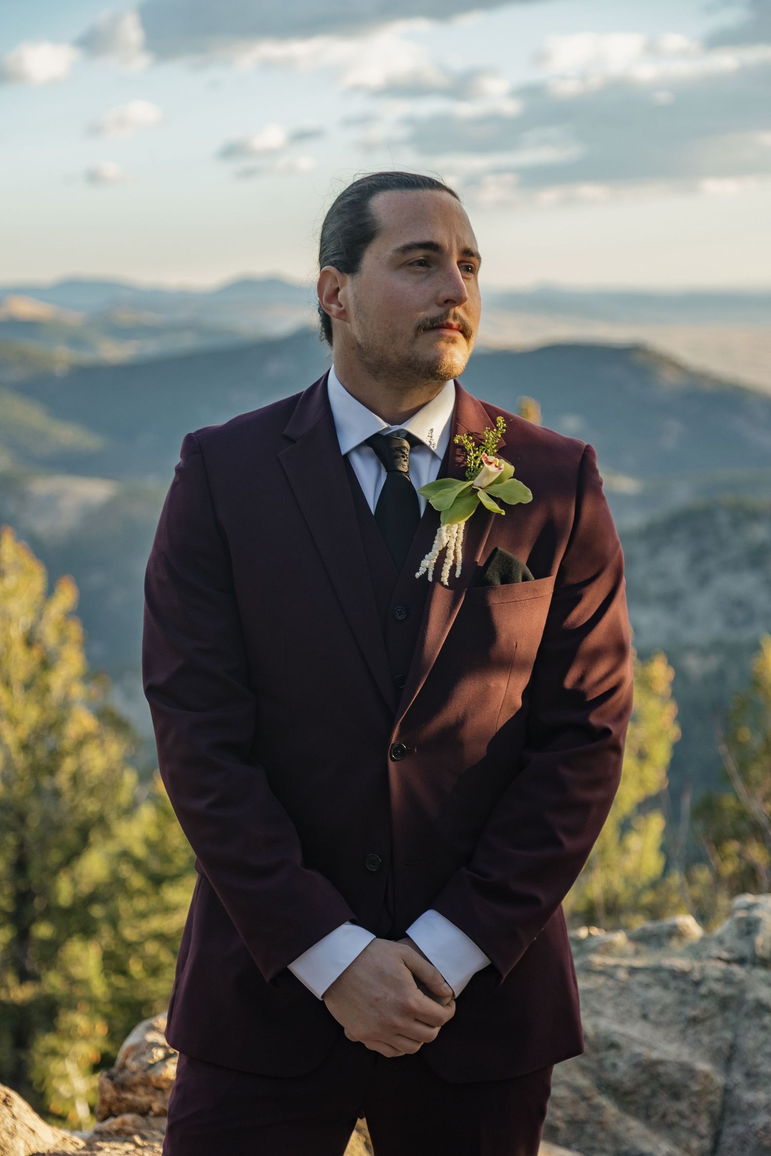 Groom in maroon three-piece suit with floral boutonniere stands at mountain vista.