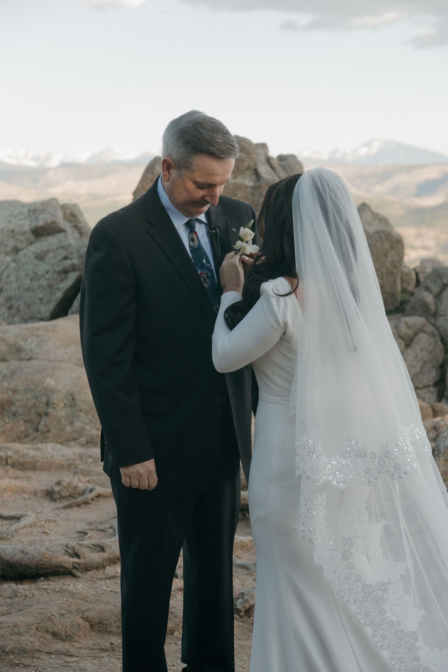 Groom in black suit holds bride's hand during outdoor wedding ceremony at rocky mountain location.