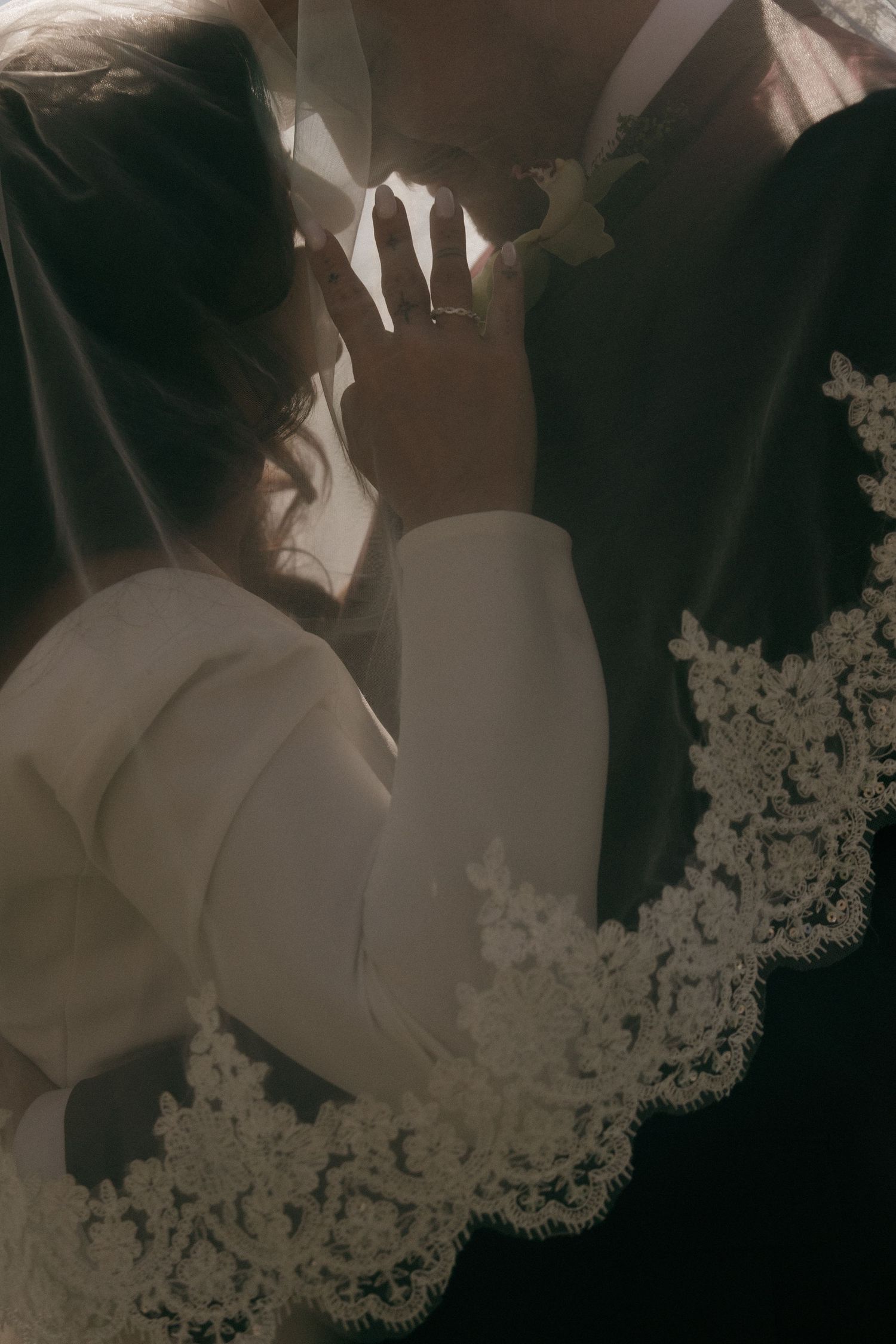 Romantic close-up of bride and groom nearly kissing beneath lace-edged veil with wedding ring visible.