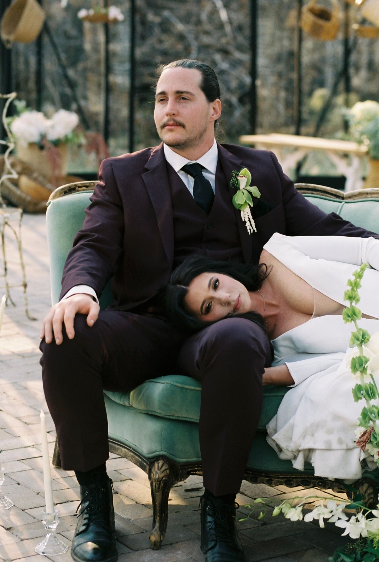Bride rests her head on groom's lap as they sit on a vintage green velvet sofa at their wedding.