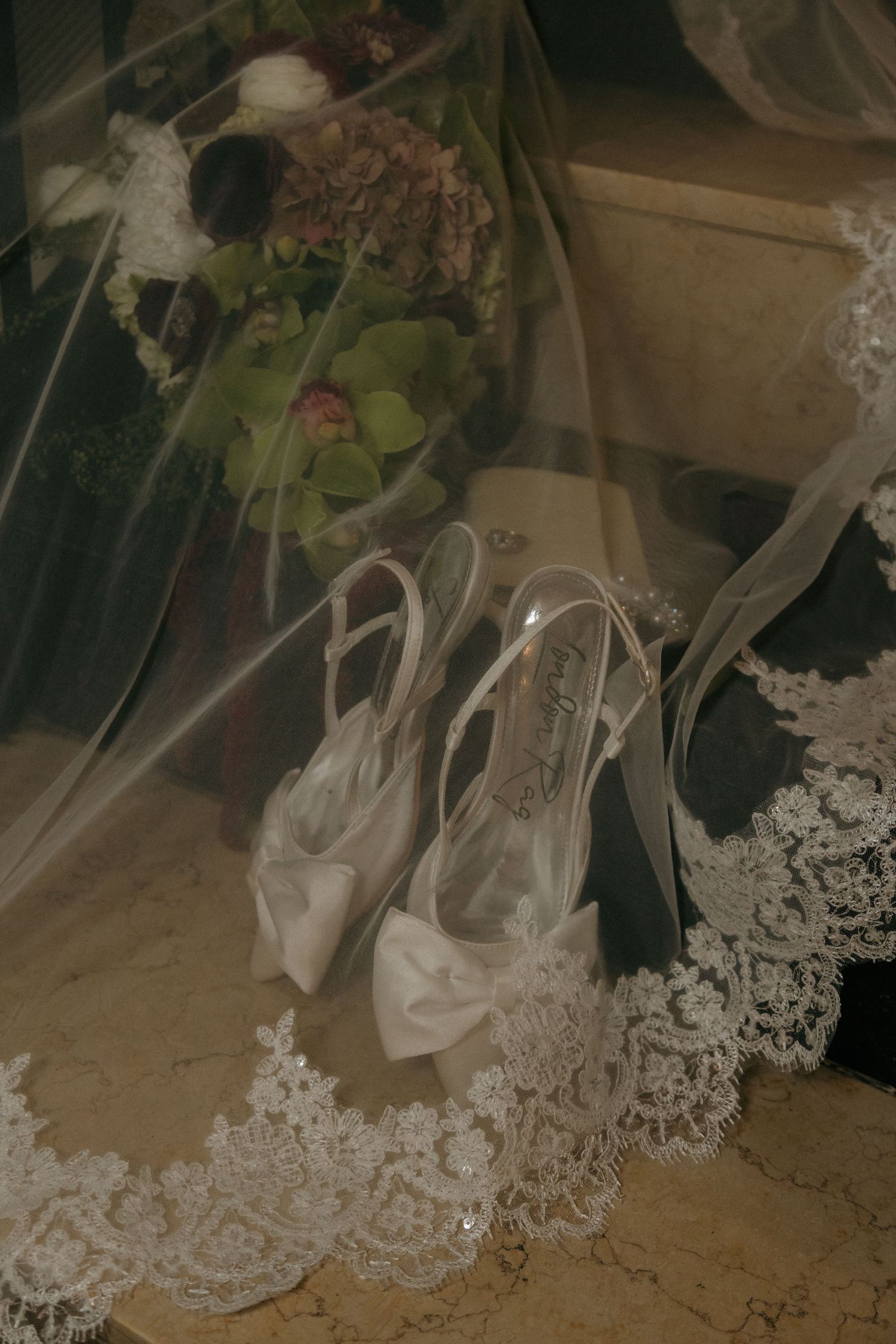 Bridal white satin heels with bow detail rest beside a floral bouquet under a lace-trimmed wedding veil.