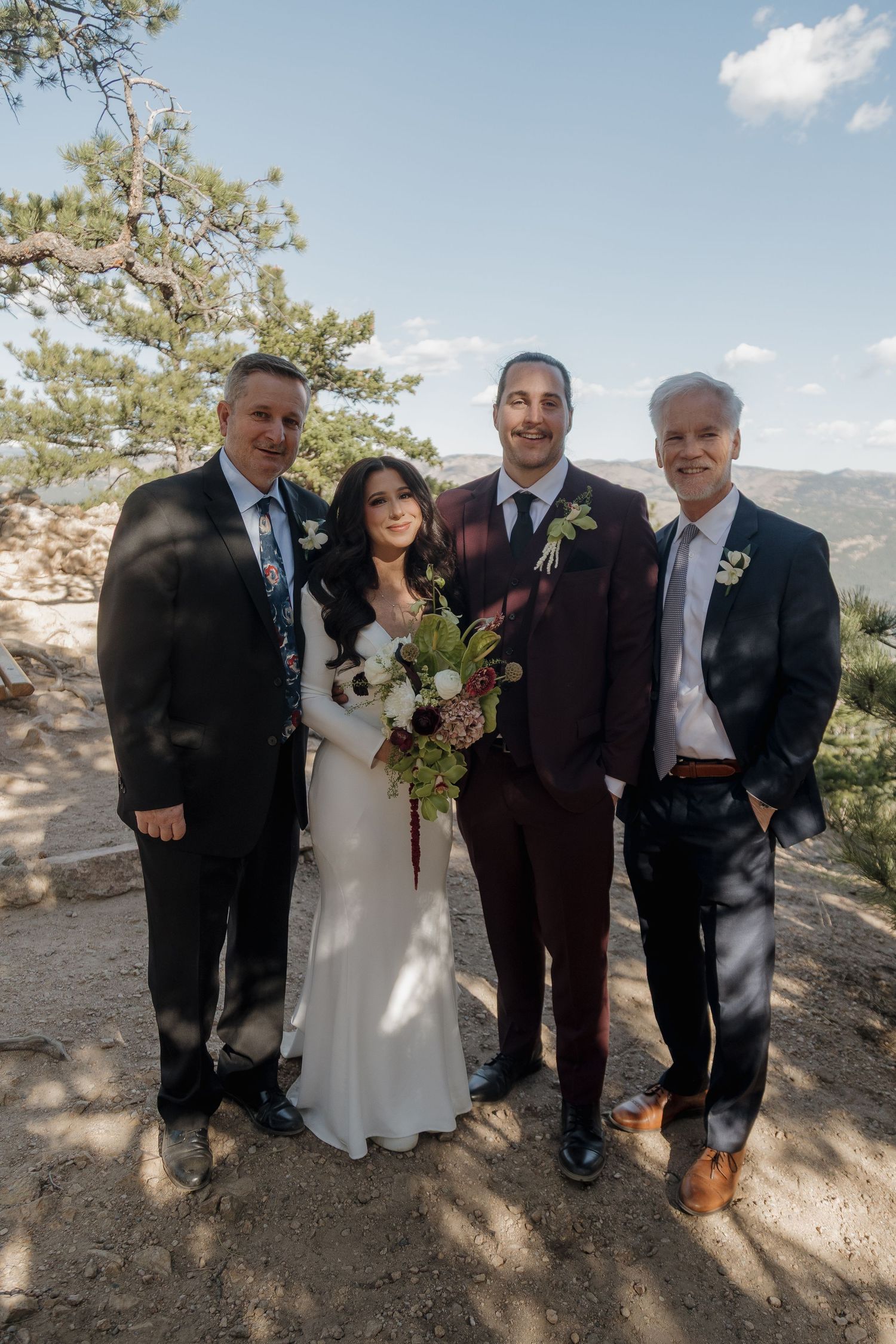 Bride and groom pose with two family members outdoors with mountain views on their wedding day.