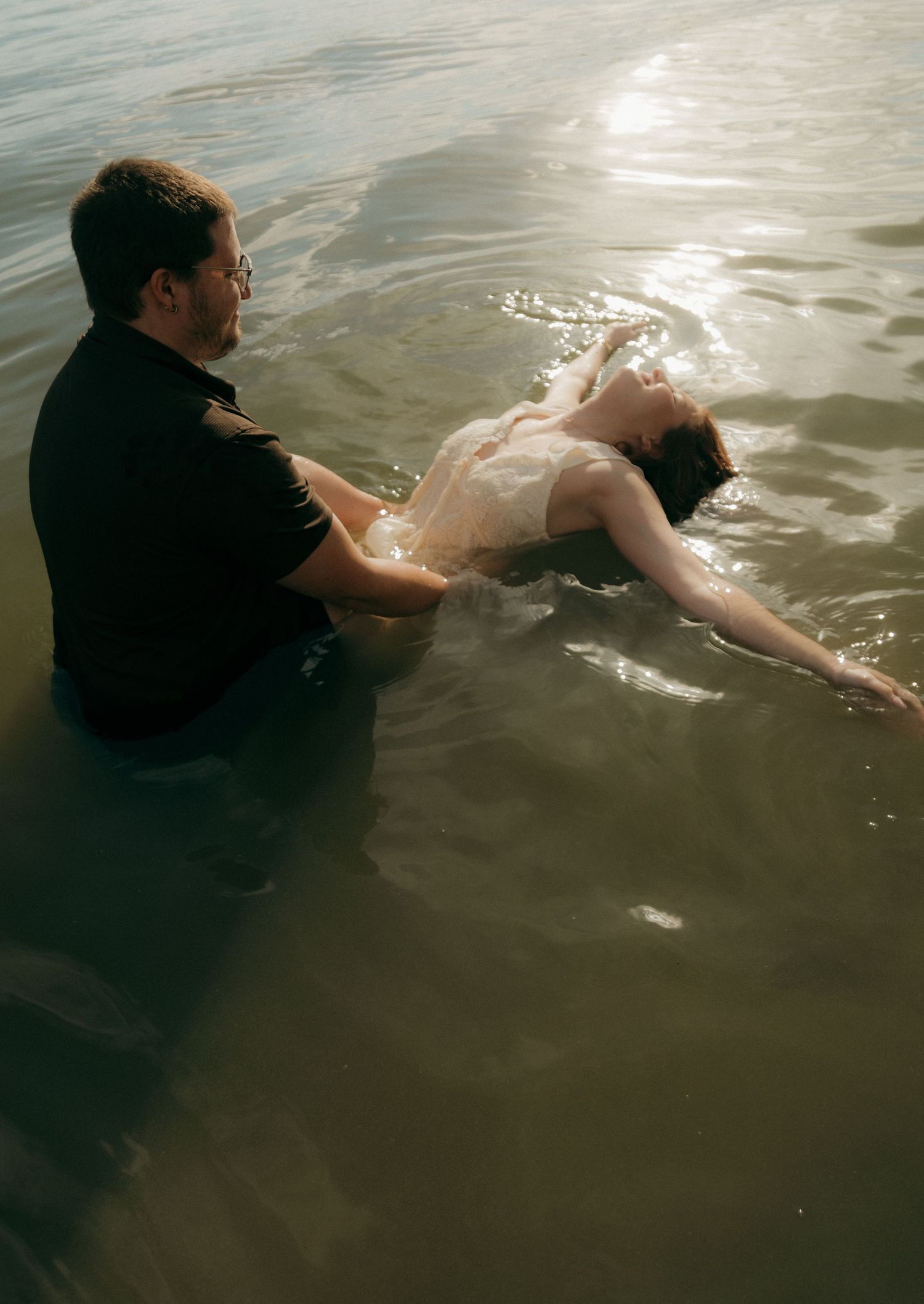 Couple in wedding attire sitting in shallow water, bride floating back as groom supports her in golden light.