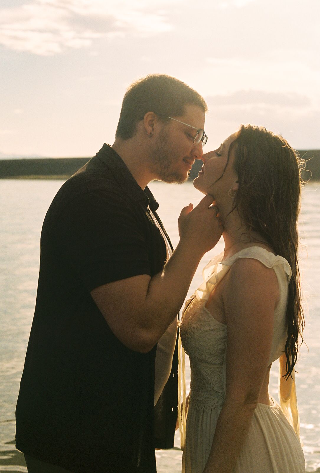 Couple nearly kissing in a tender moment by a sunlit lake, backlit by golden hour light.
