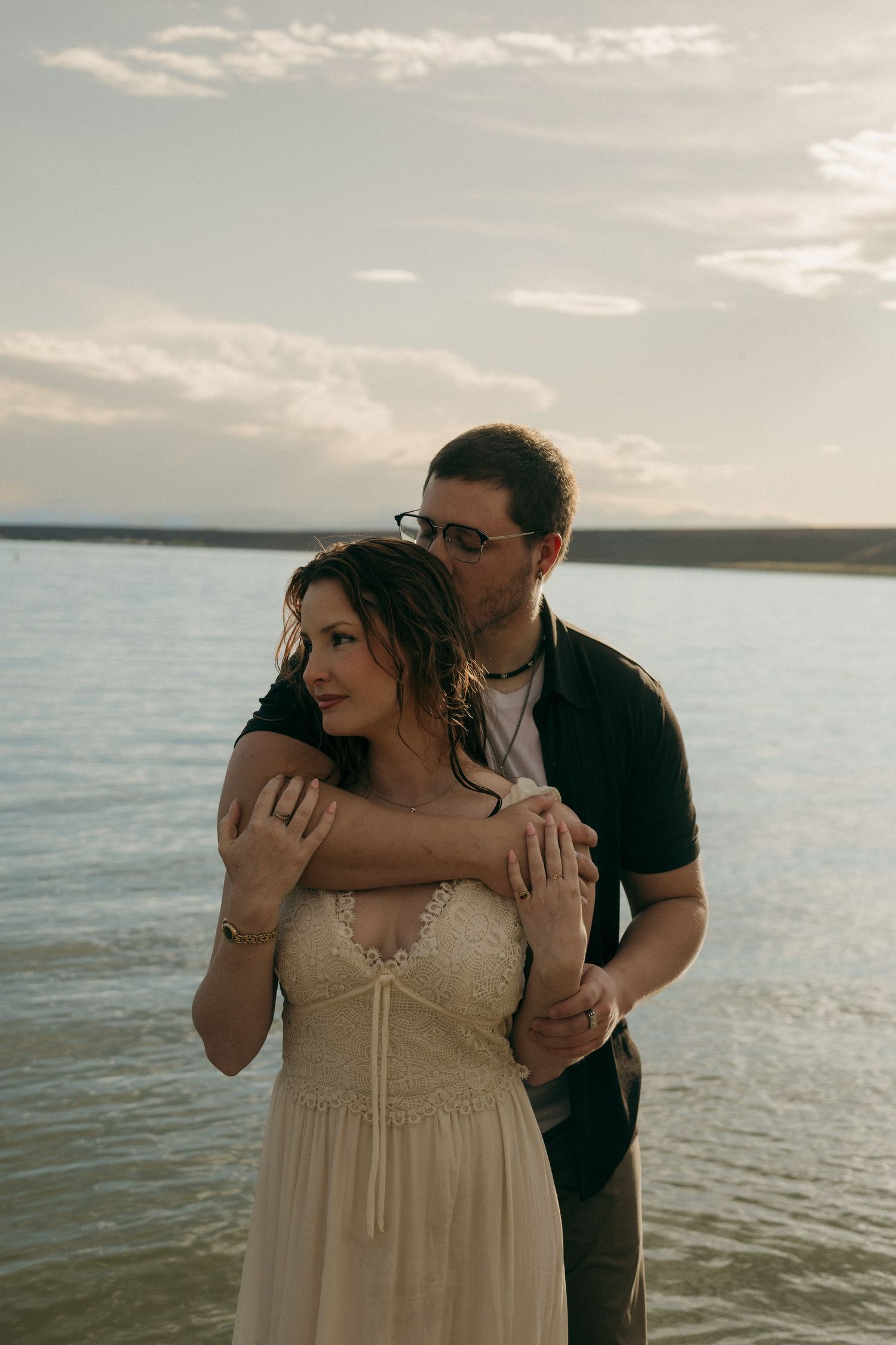 Couple embracing by a lake at dusk, man in black shirt holding woman in white lace dress from behind.