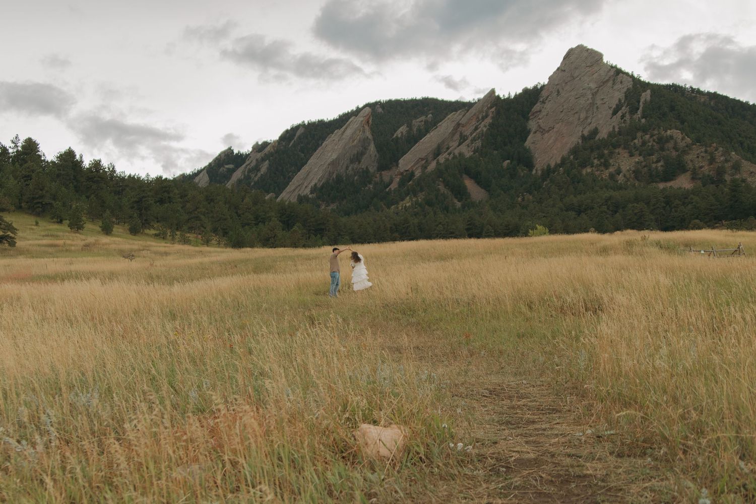 A couple holds hands while walking through a vast golden field with the Flatirons in the distance.