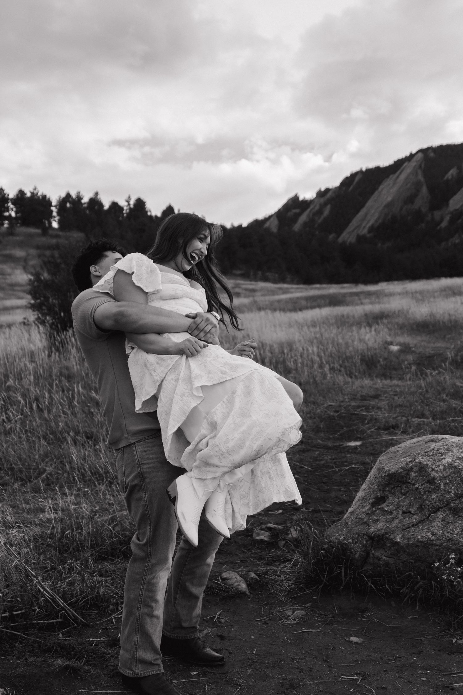 Black and white photo of man lifting woman in white dress in mountain meadow.