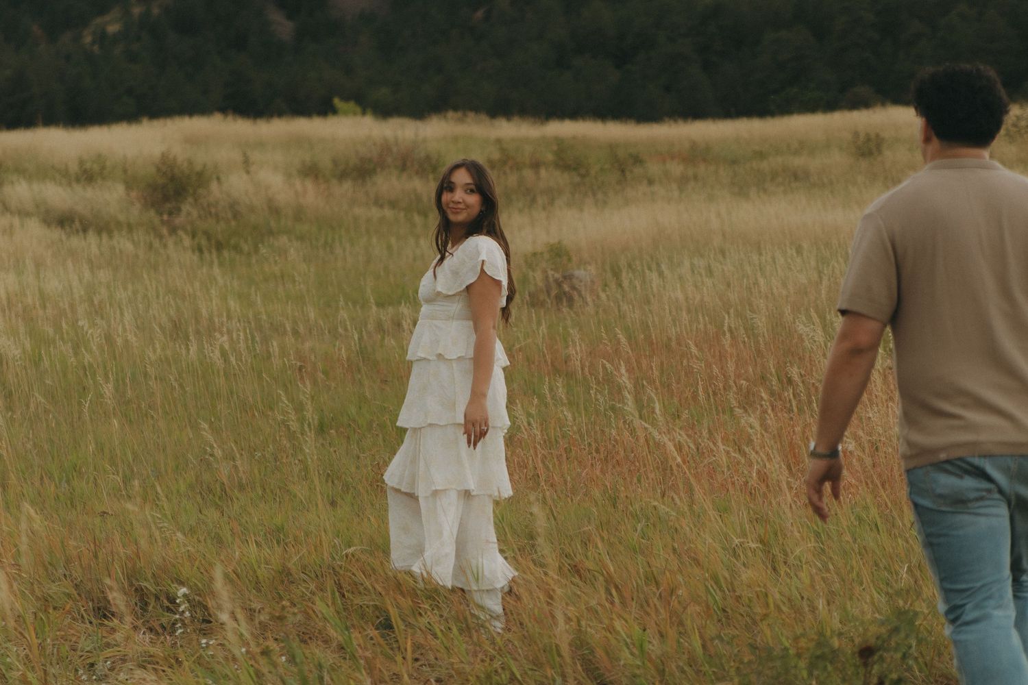 Woman in white tiered dress smiling in golden grass field with man walking toward her.