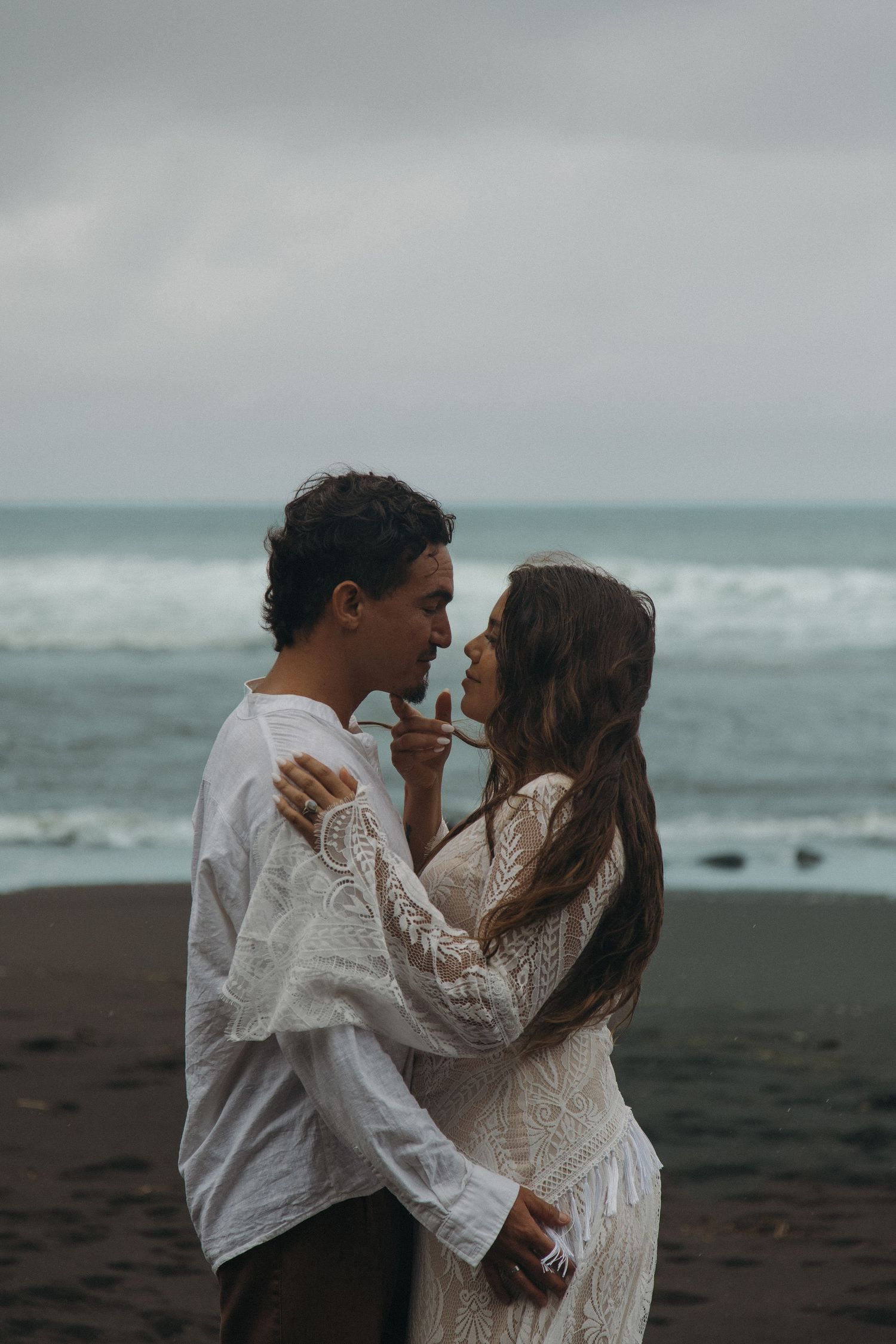 Couple in white outfits sharing an intimate moment on a dark sandy beach with ocean waves behind them.