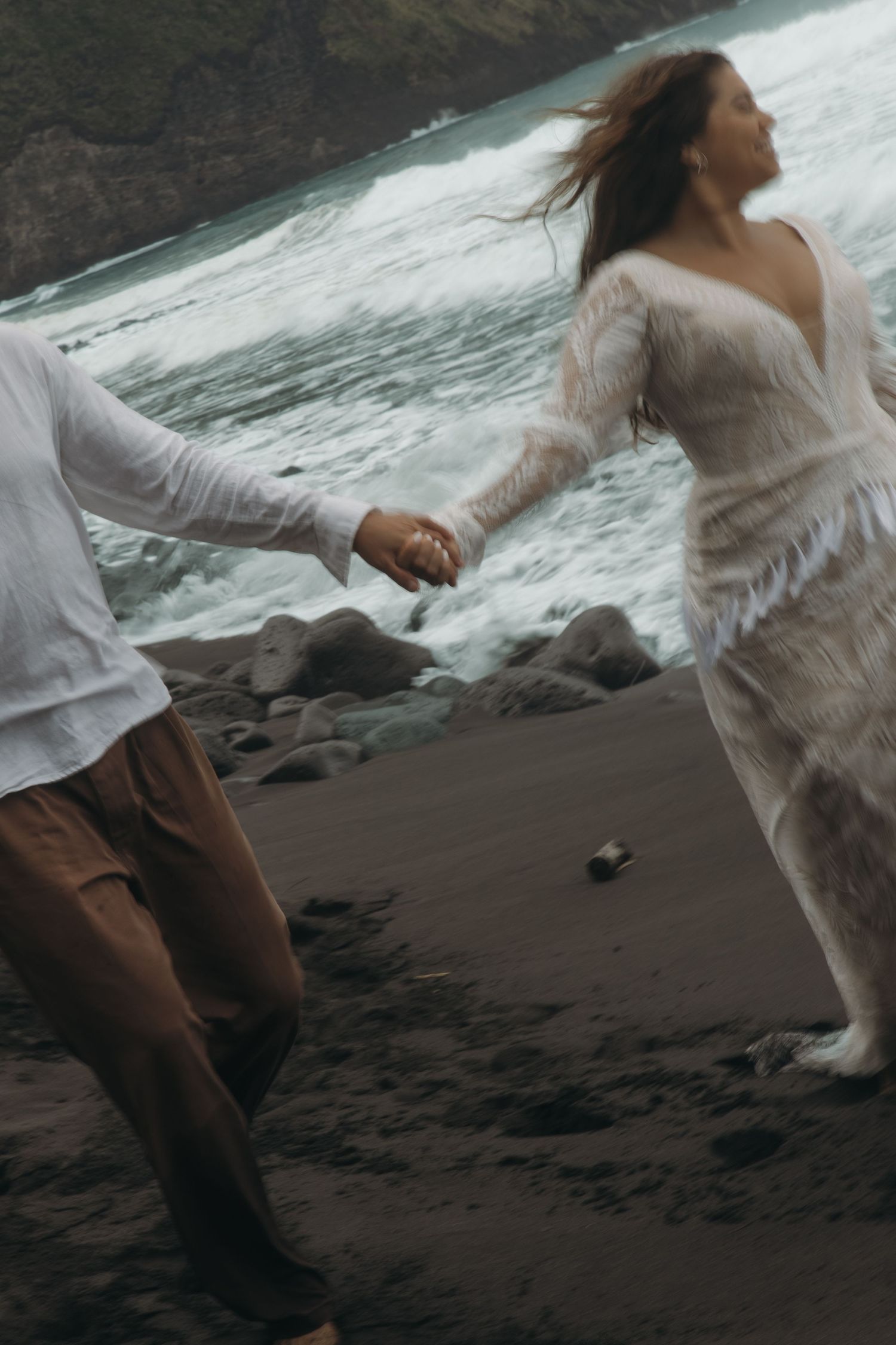 Couple runs hand-in-hand along black sand beach with dramatic ocean waves crashing behind them.
