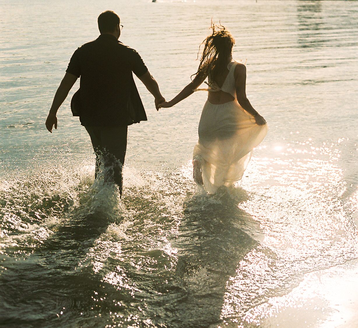 Couple holding hands running through ocean waves at sunset in wedding attire, backlit by golden light.