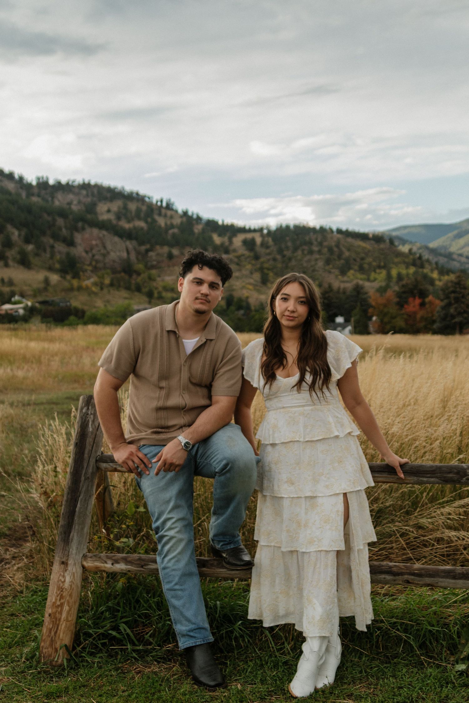 Couple sitting on rustic wooden fence with scenic mountain and golden field backdrop.
