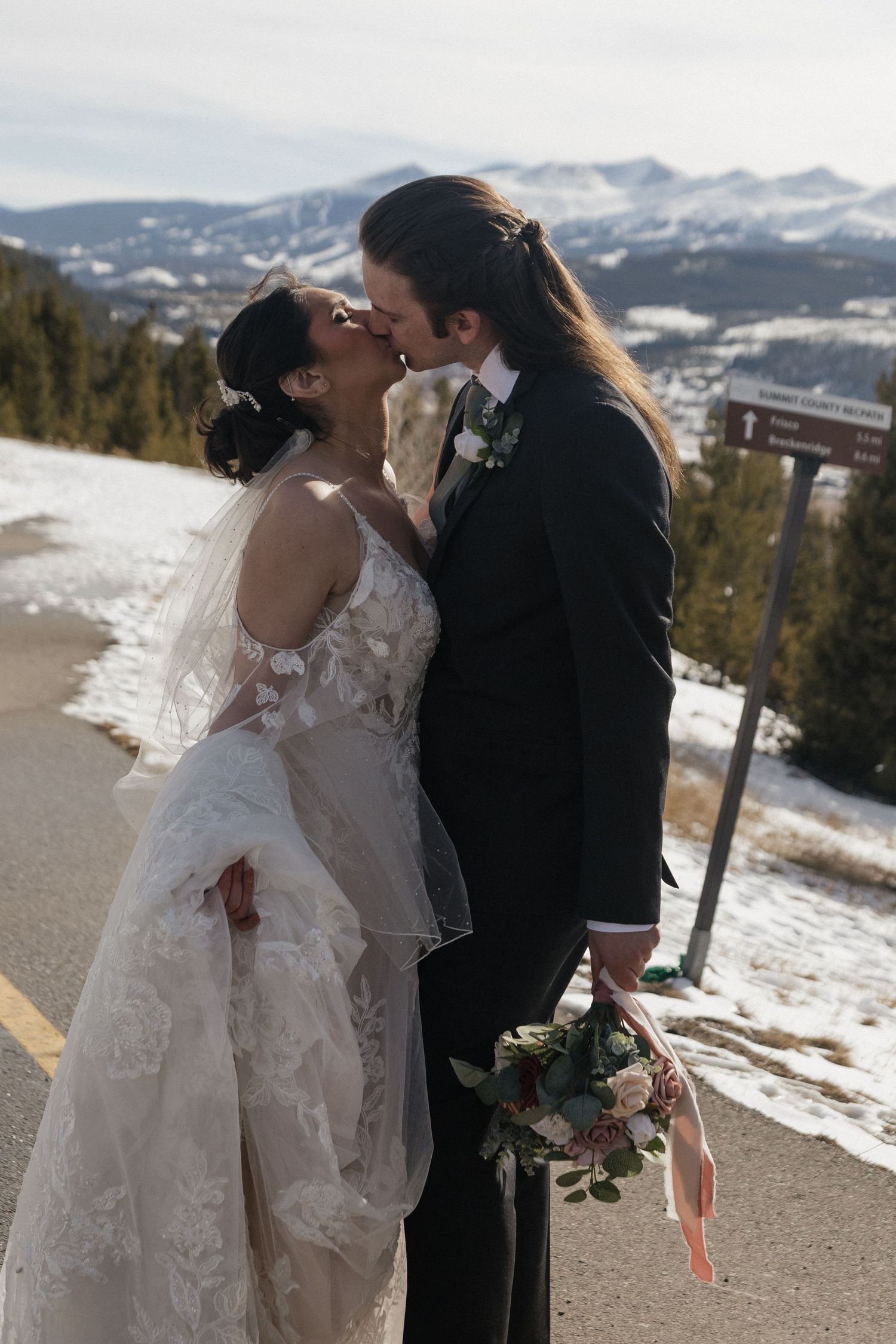 A winter wedding couple embraces on a snow-covered mountain overlook with scenic mountain vistas in the background.