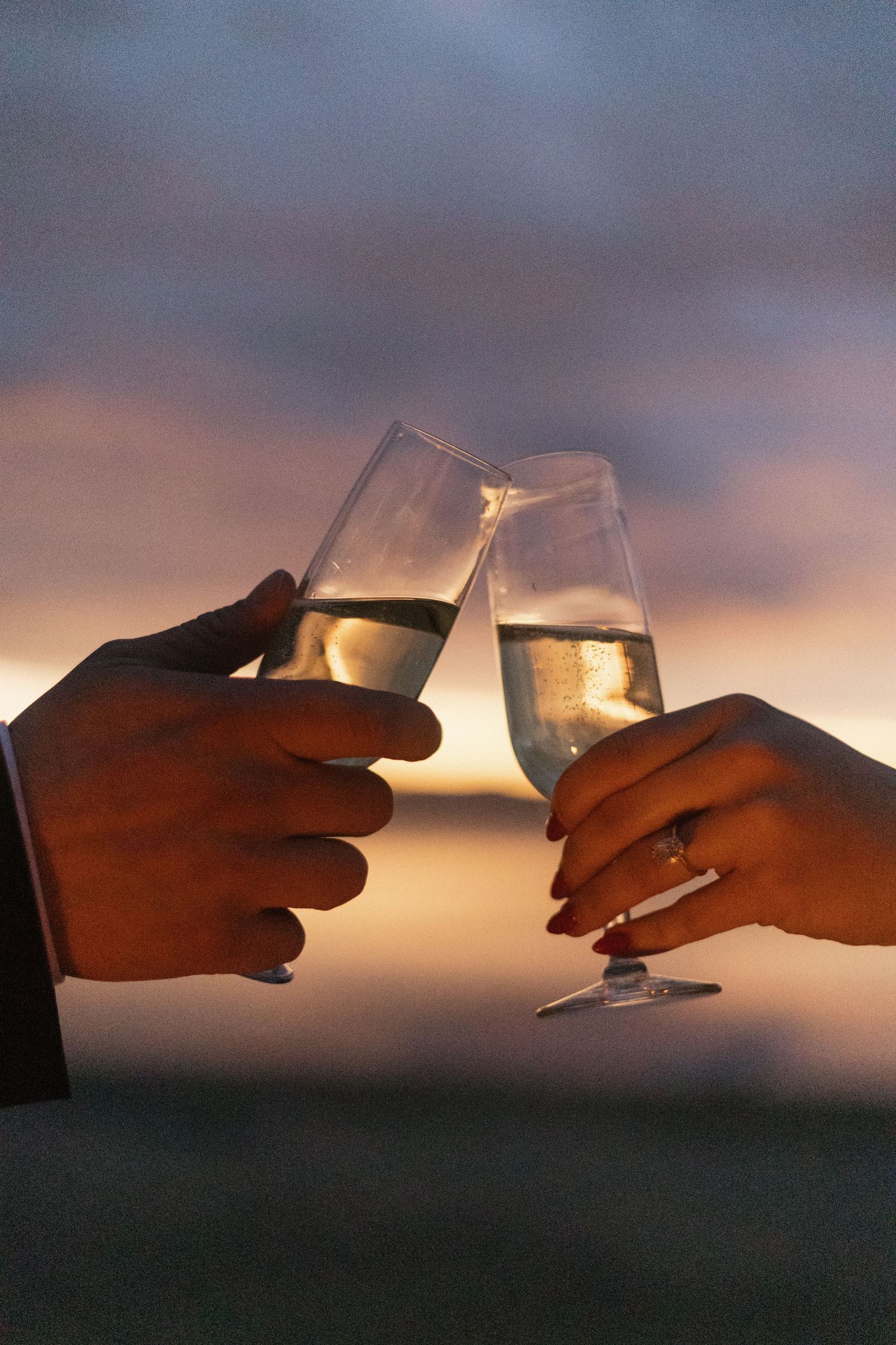 Two hands toast wine glasses against a dramatic sunset sky with silhouetted mountains in the background.