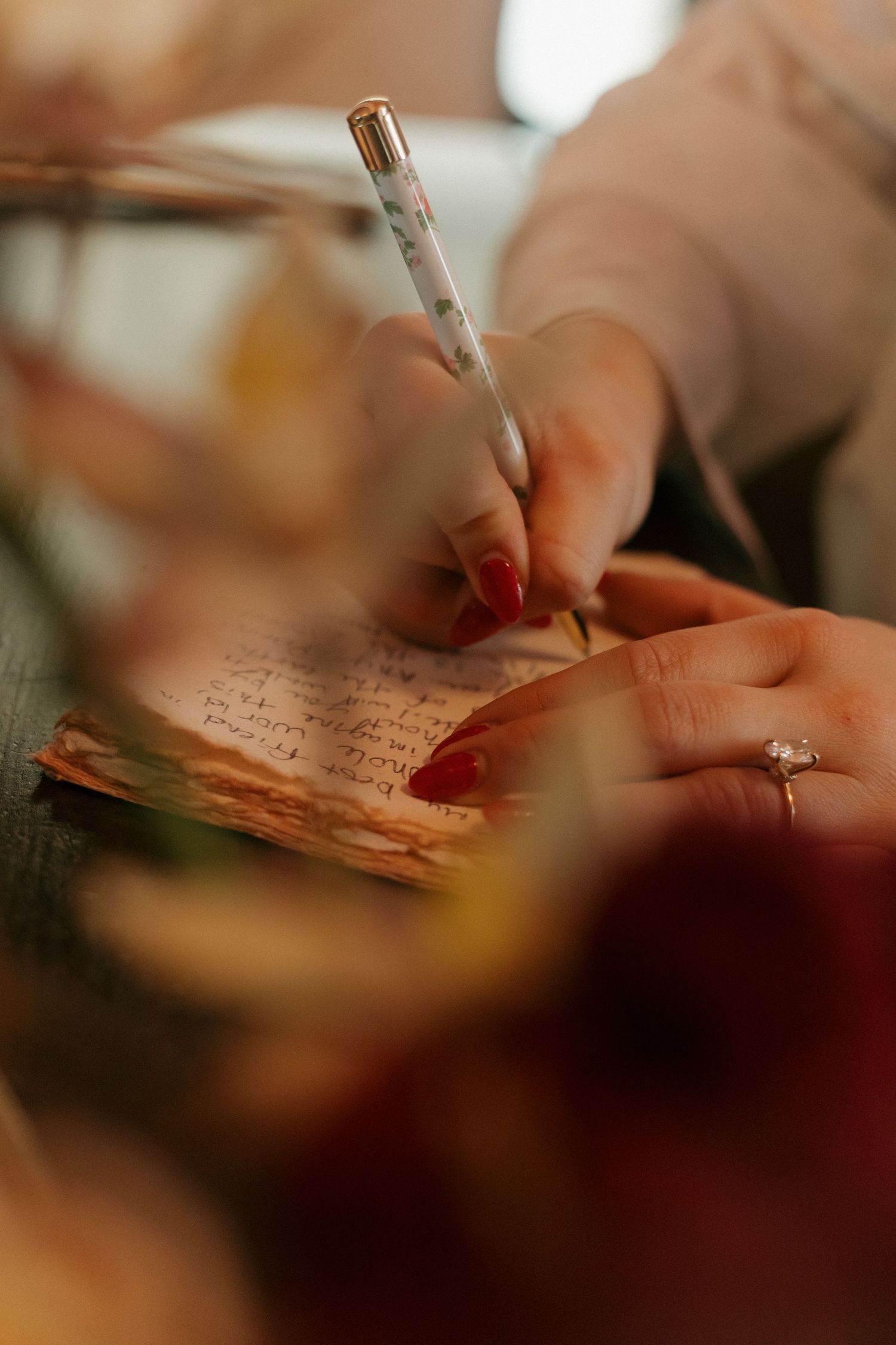 A hand with red nail polish writes in a journal with a pen while wearing an engagement ring.