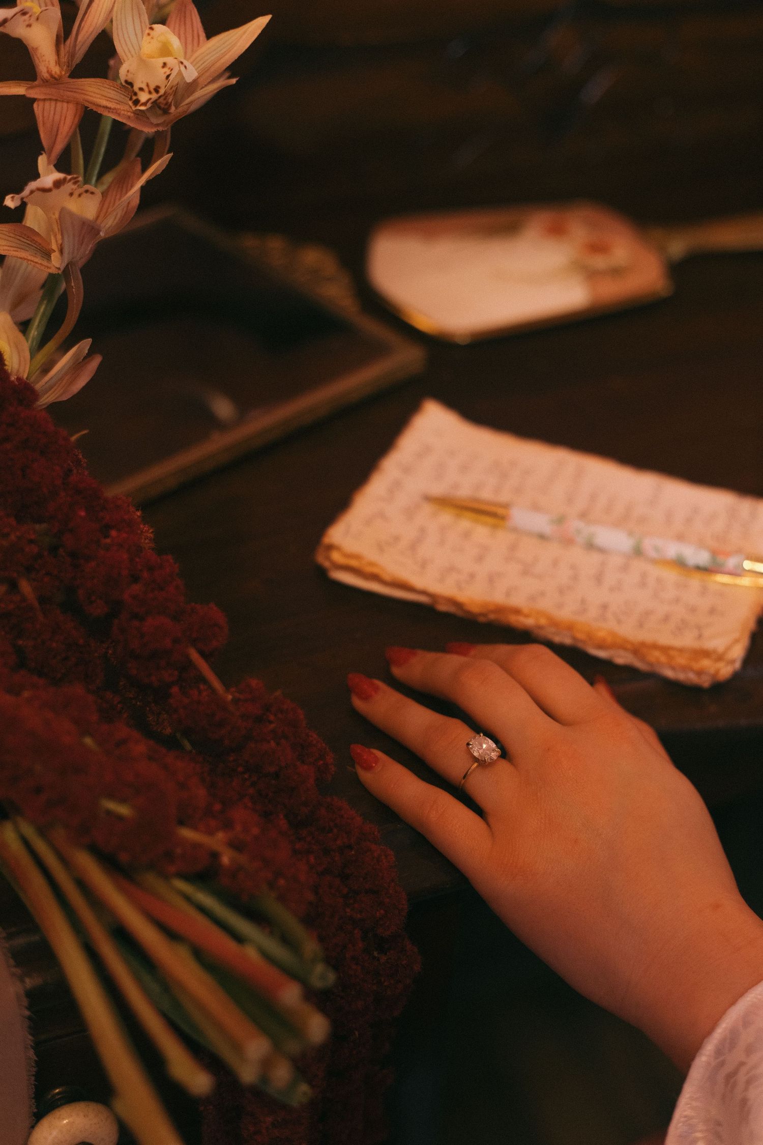 Close-up of an engagement ring on a hand next to flowers and a notebook.
