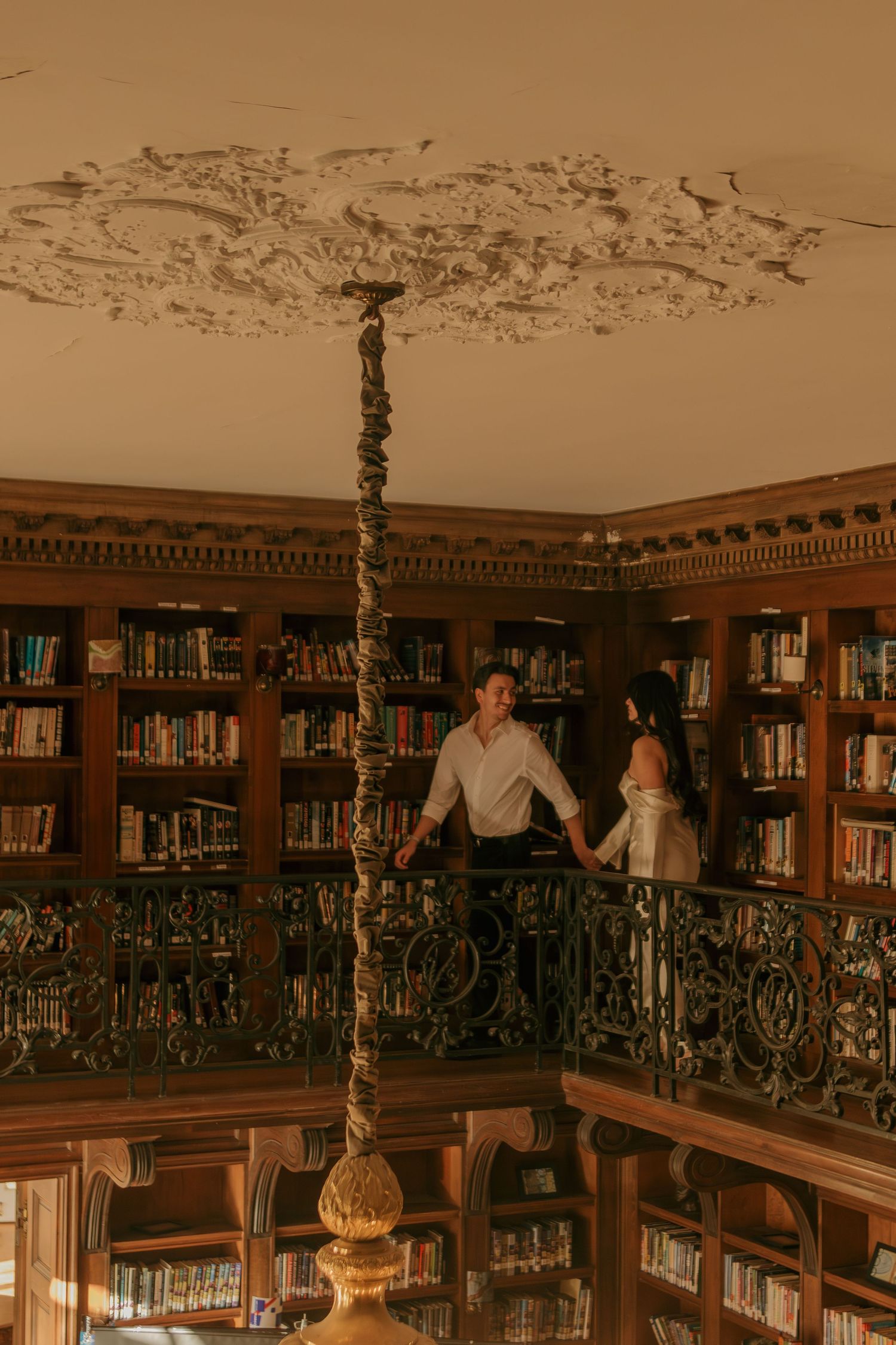 Couple stands on ornate balcony overlooking classic library interior with grand chandelier.