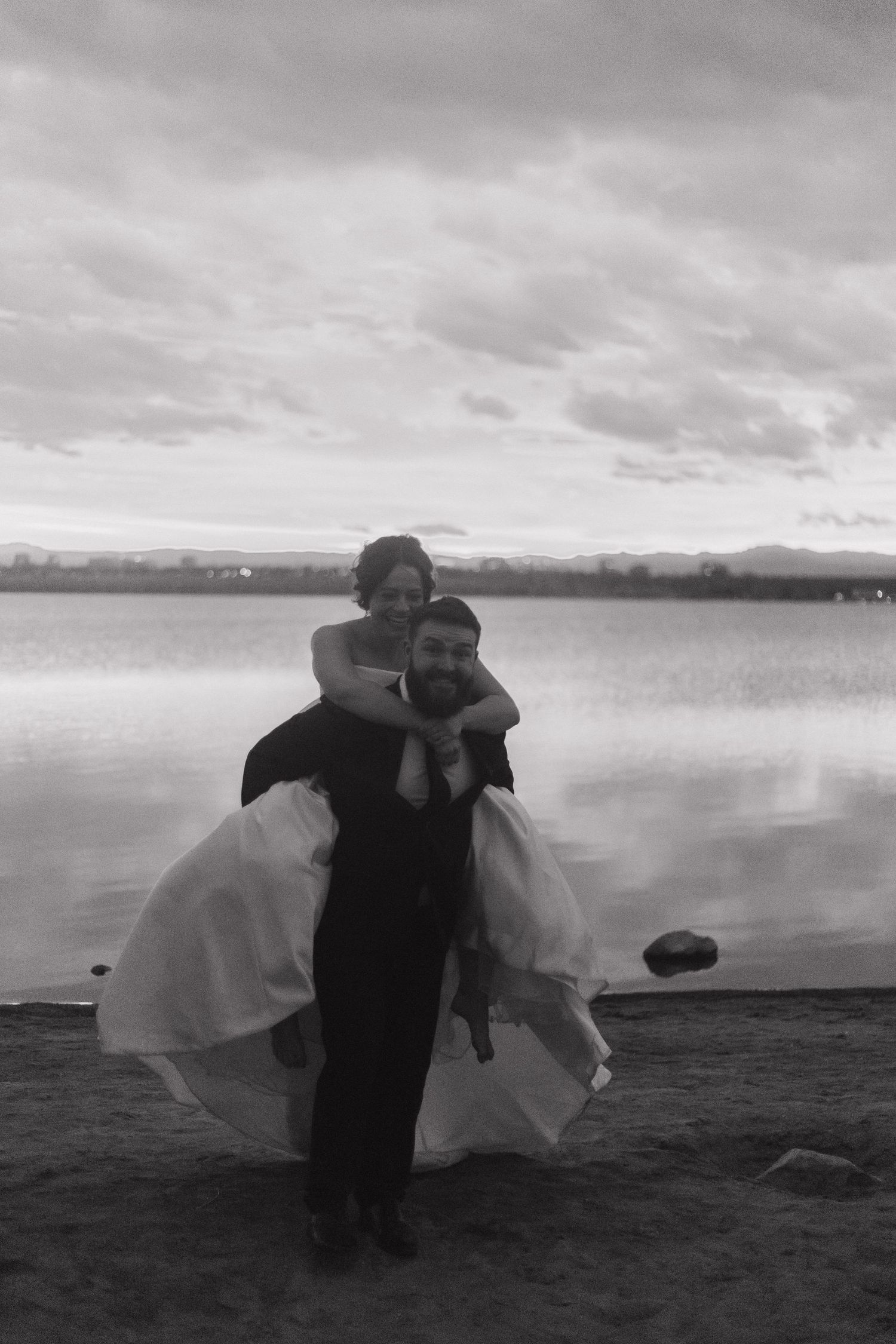 A romantic couple in wedding attire embraces on a beach with cloudy skies in black and white.