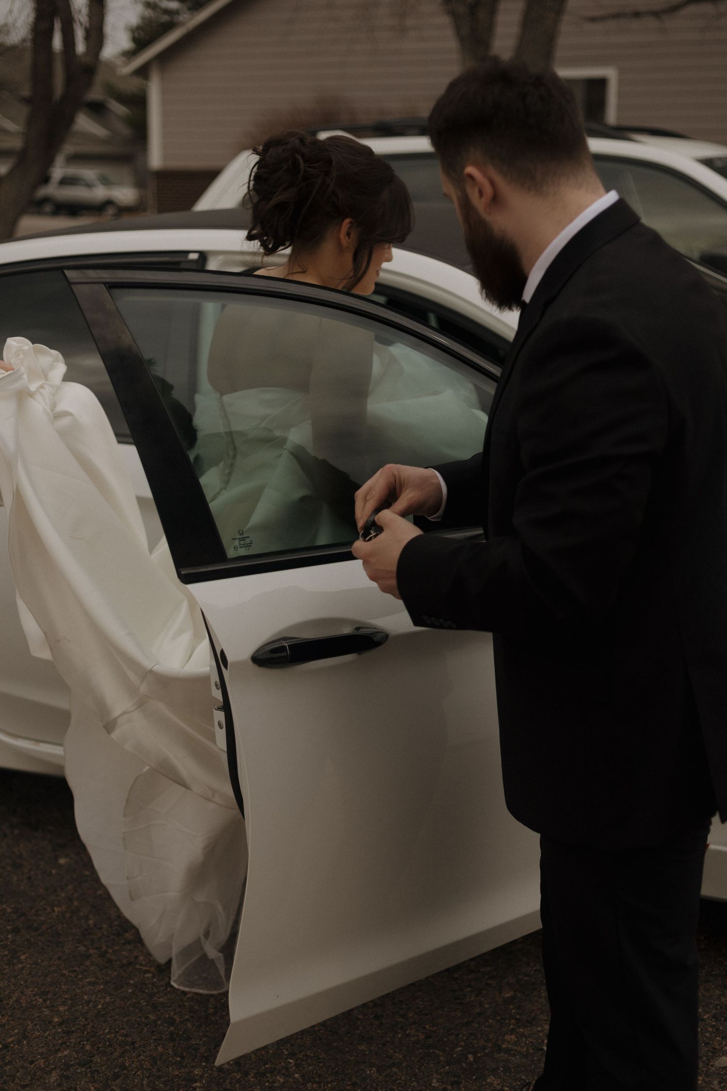 A groom helps his bride into a white car after their wedding ceremony.