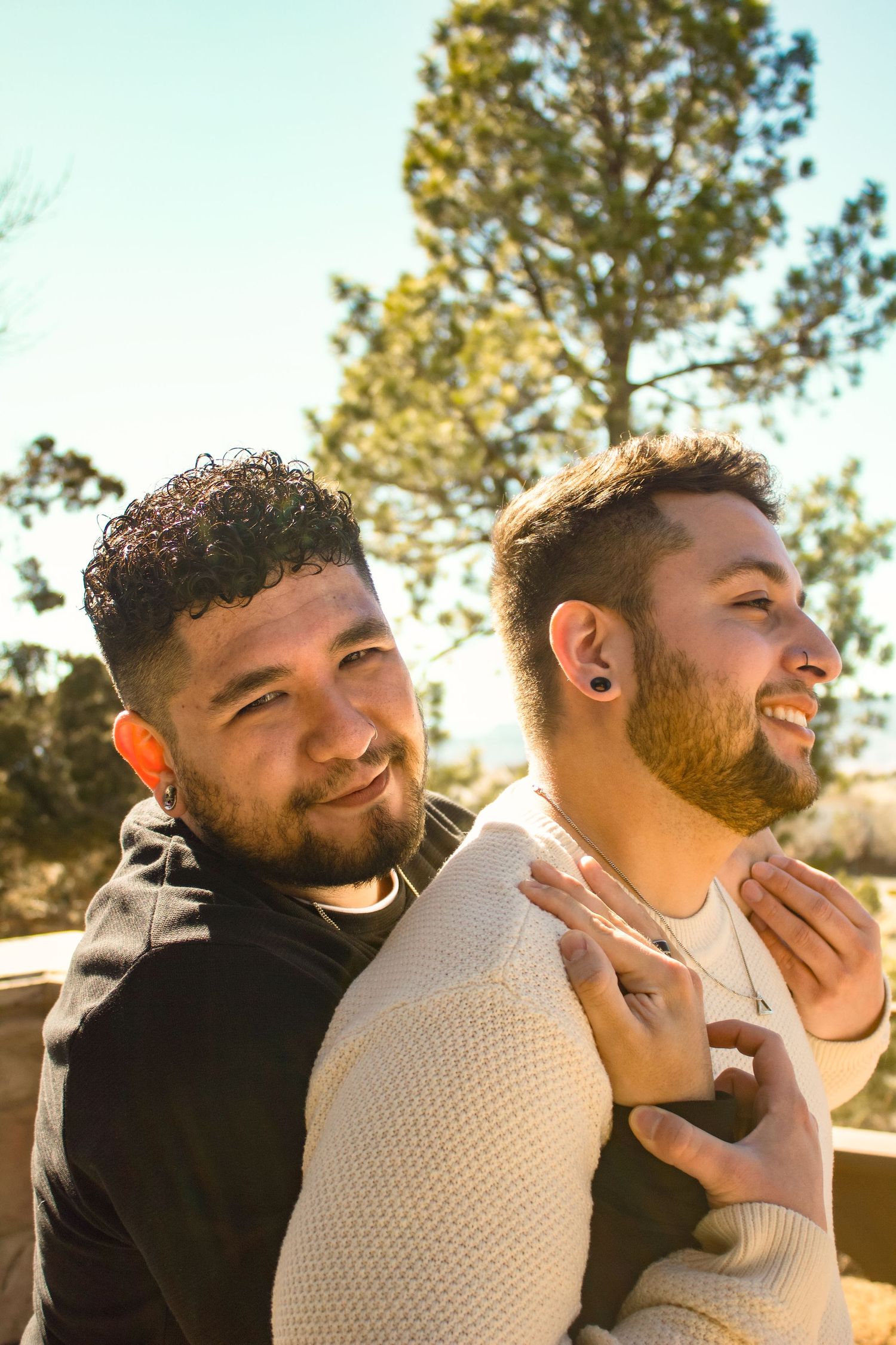 Two people laugh and embrace outdoors with pine trees in the background during golden hour.