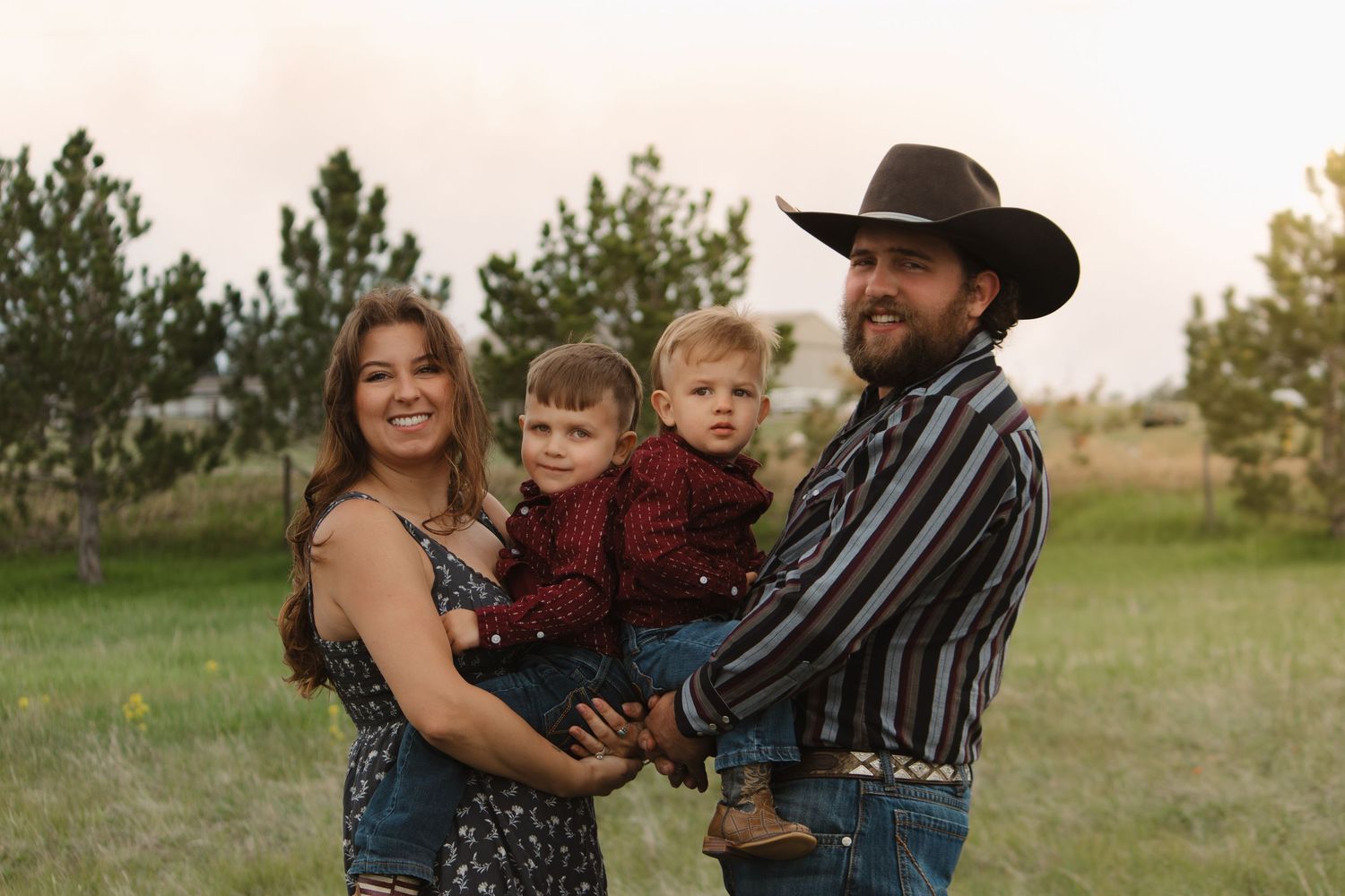 A western-styled outdoor family portrait in a grassy field with pine trees at sunset.
