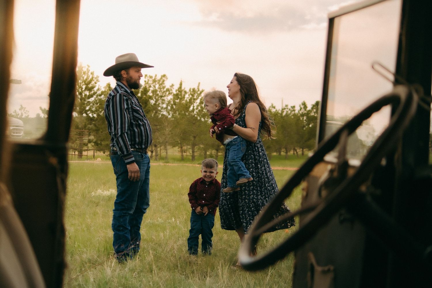 Through an old truck window, a family shares a candid moment in a rural field at dusk.
