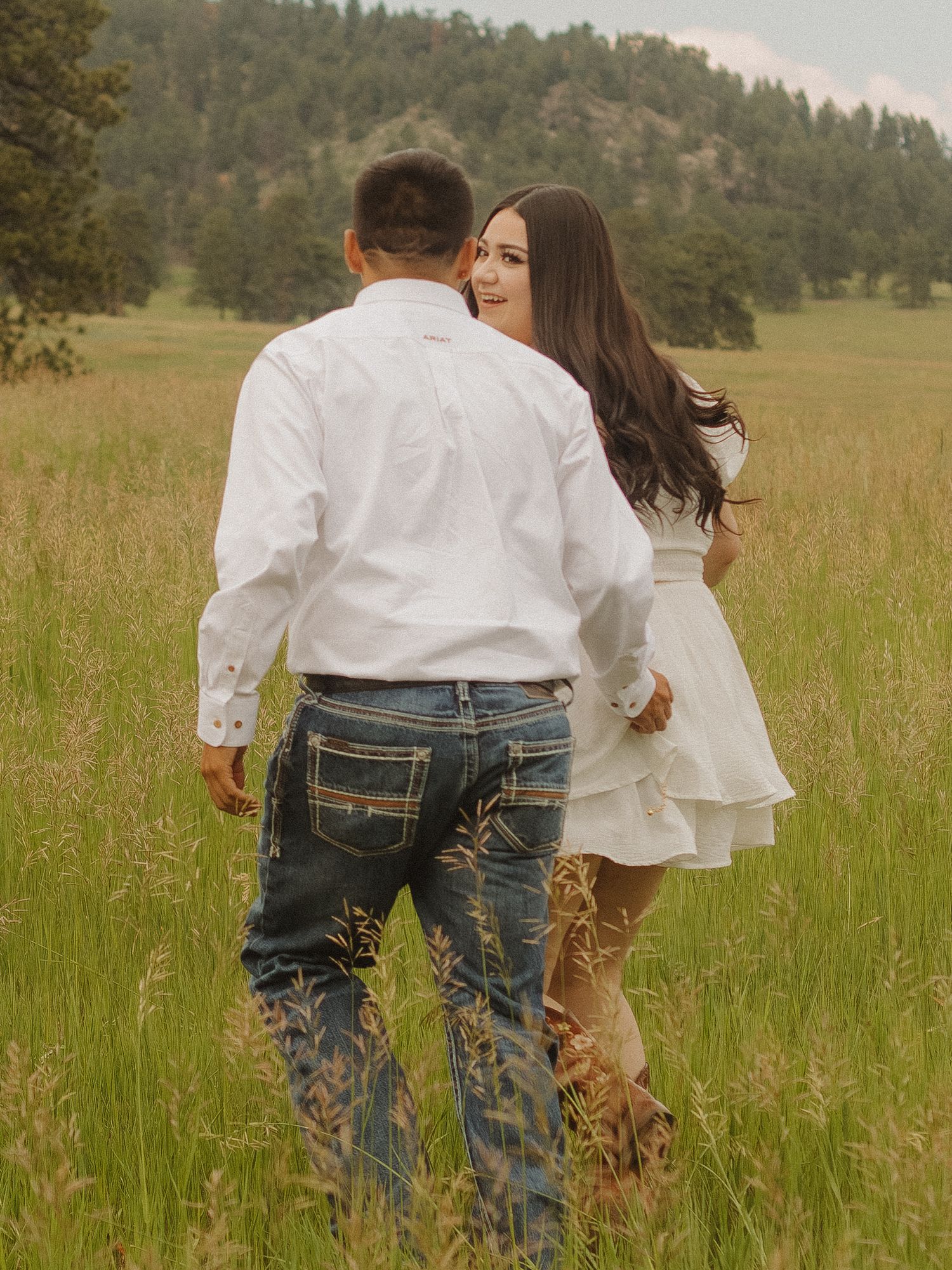 Couple sharing an intimate moment while walking through tall grass with mountains in background.