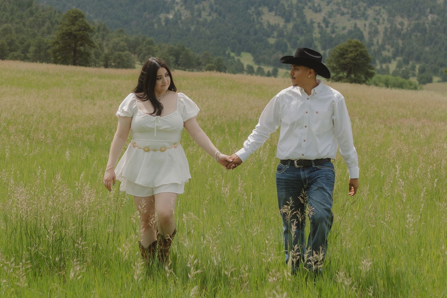 Couple in western attire walking hand in hand through mountain meadow with tall grass.