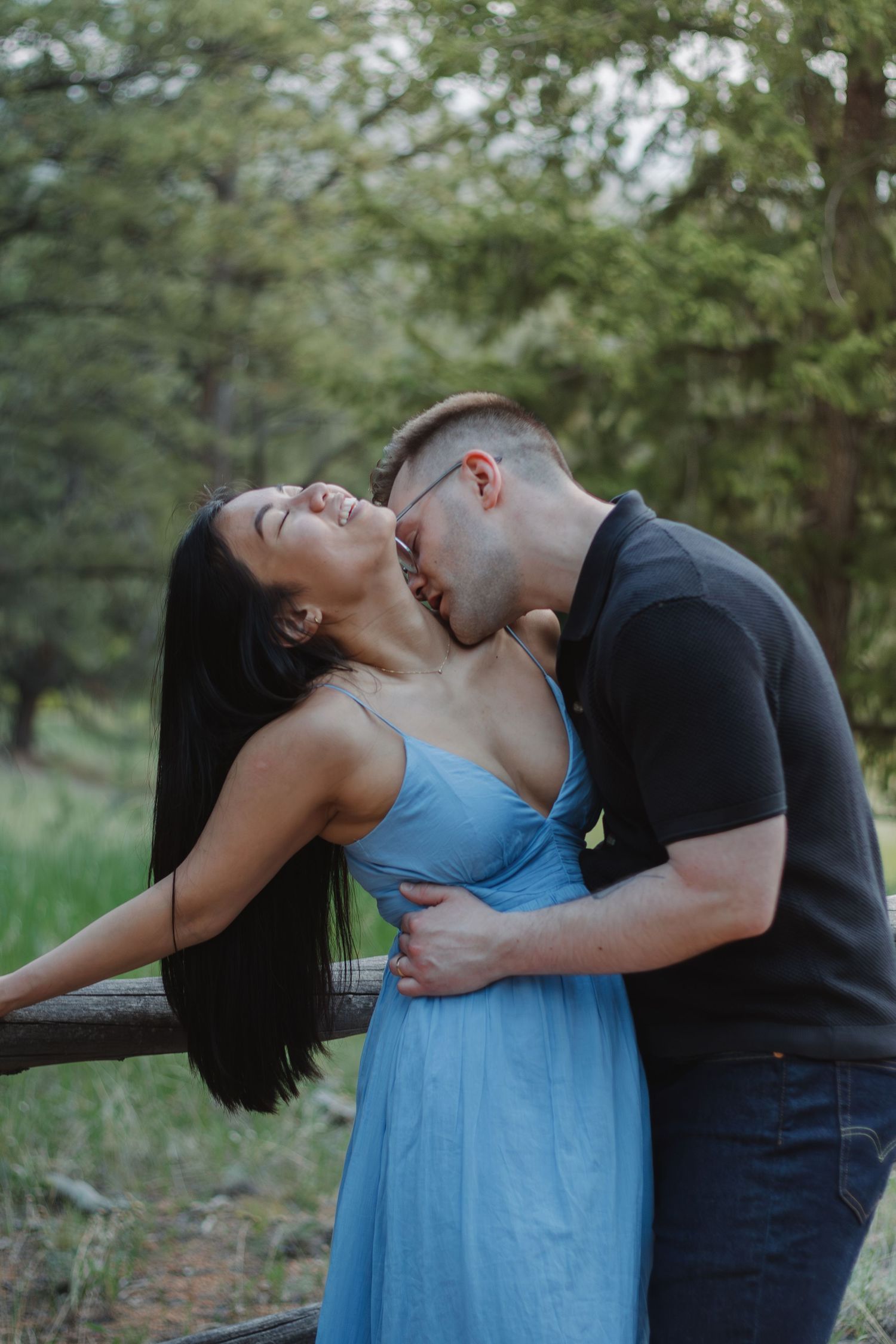 A romantic moment shared between a couple by a wooden fence in a forest setting.