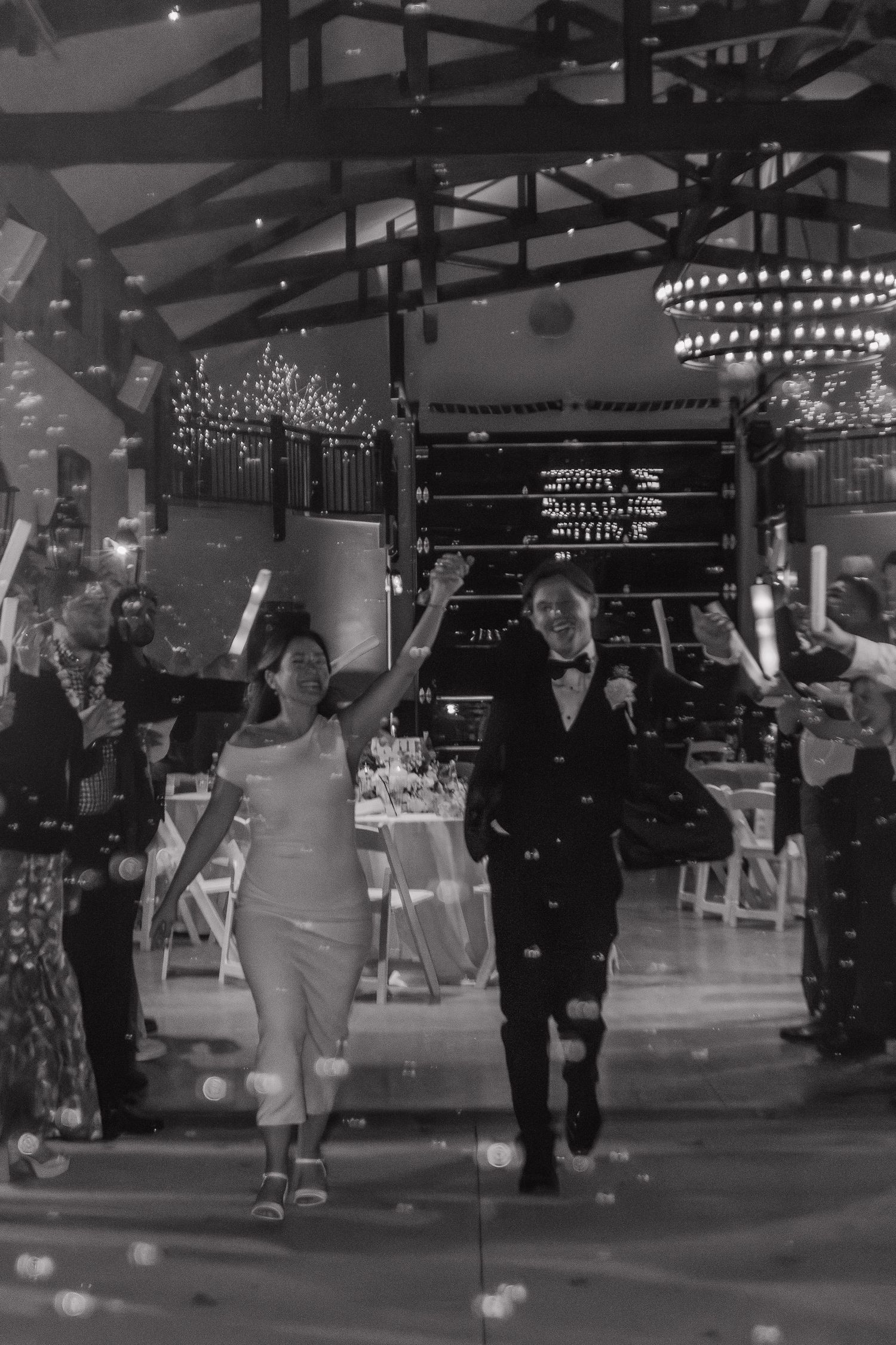 Wedding guests dancing in a venue decorated with overhead string lights and chandeliers.