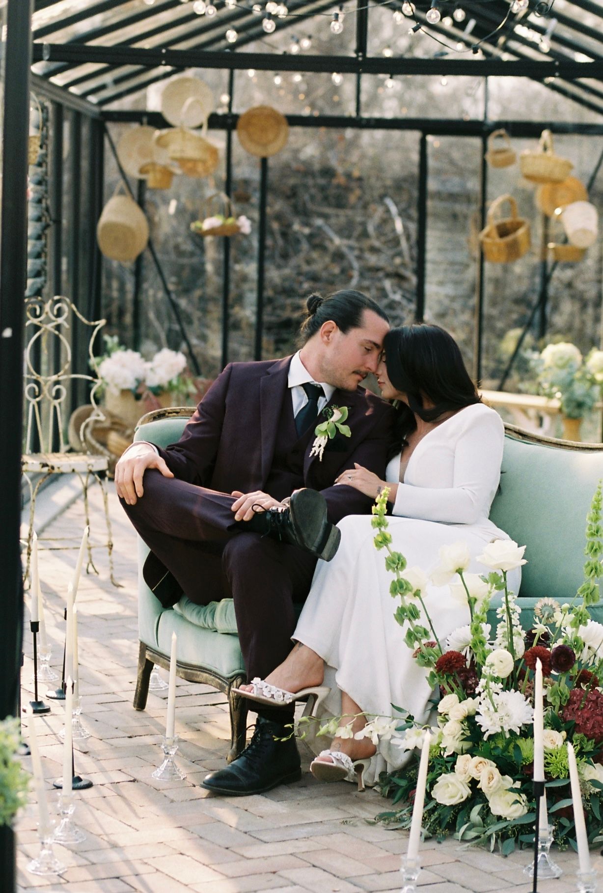 Bride and groom share intimate moment on vintage sofa inside decorated greenhouse with hanging lanterns and flowers.
